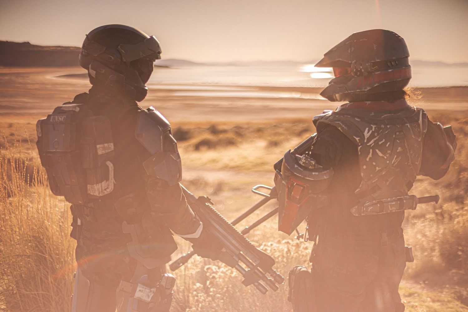 Two cosplayers in futuristic military armor stand backlit by golden sunlight near a desert lake.