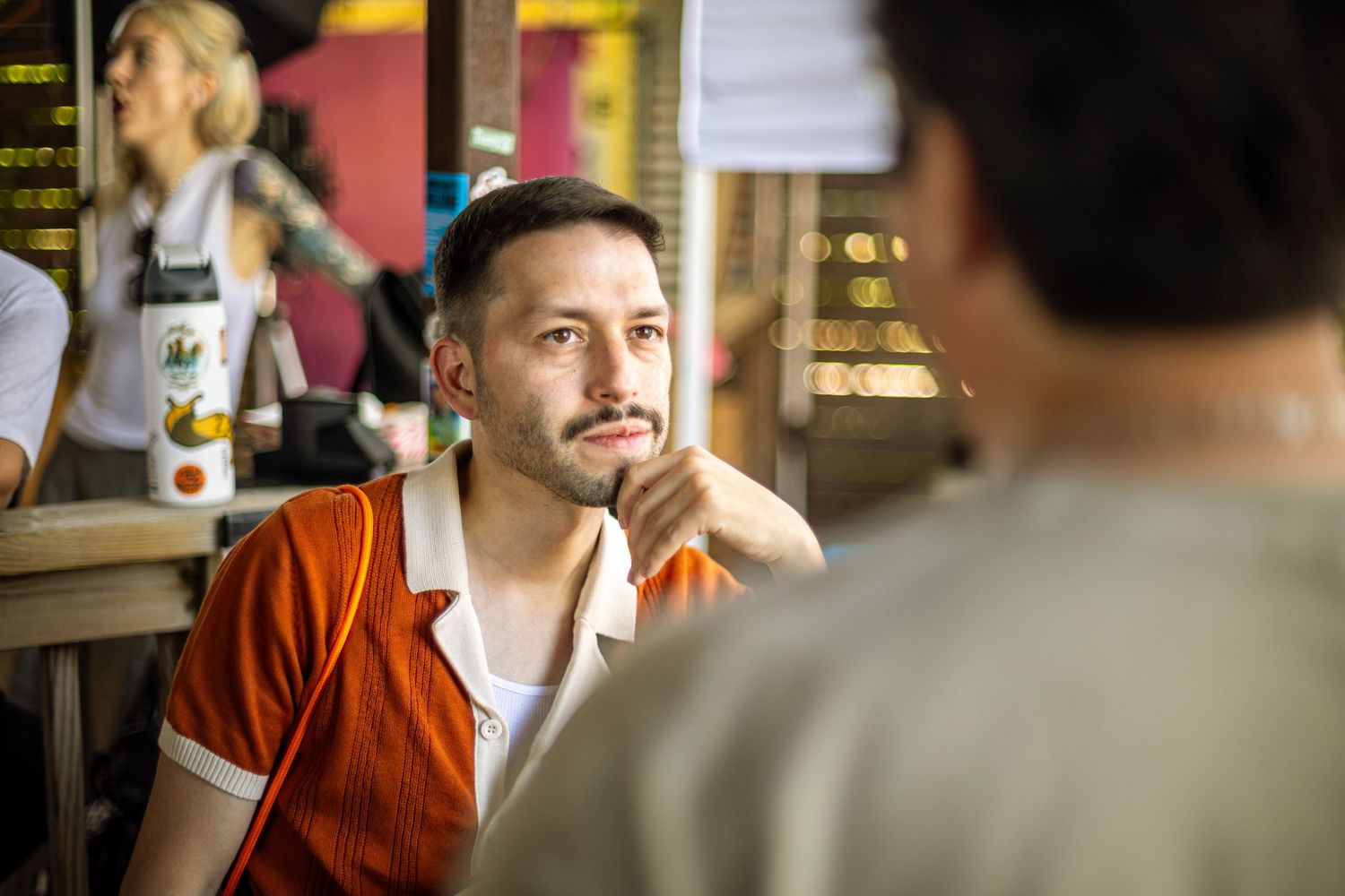 Young man in orange polo shirt sits at bar counter in conversation with another person.