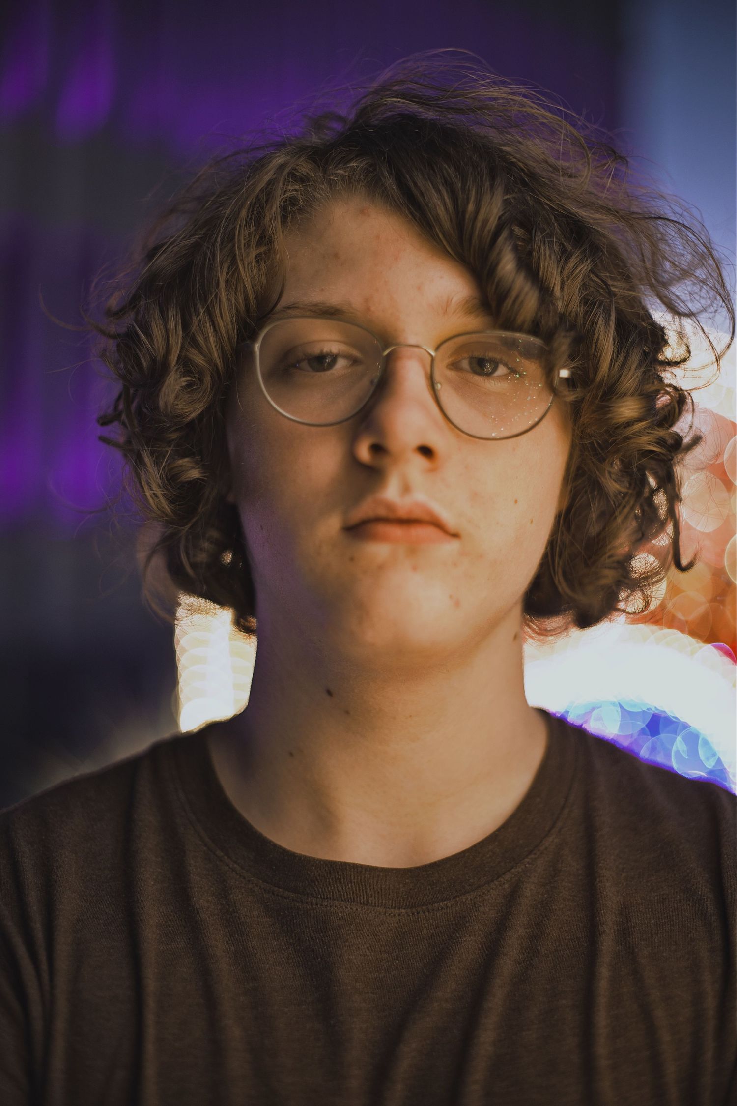 Teenage boy with curly hair and round glasses in moody neon-lit close-up portrait.