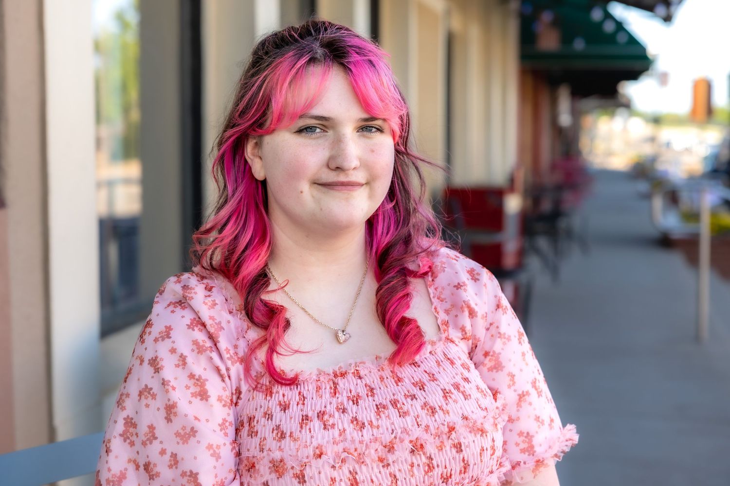 Smiling young woman with vibrant pink hair wearing pink floral dress on sunny urban sidewalk.