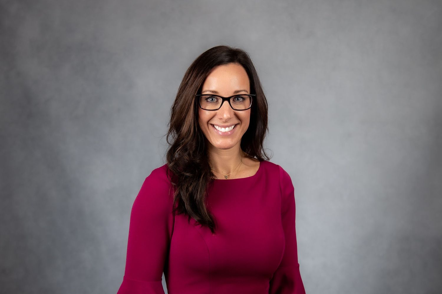 Smiling woman with long dark hair and glasses wearing a magenta top, professional headshot on gray background.