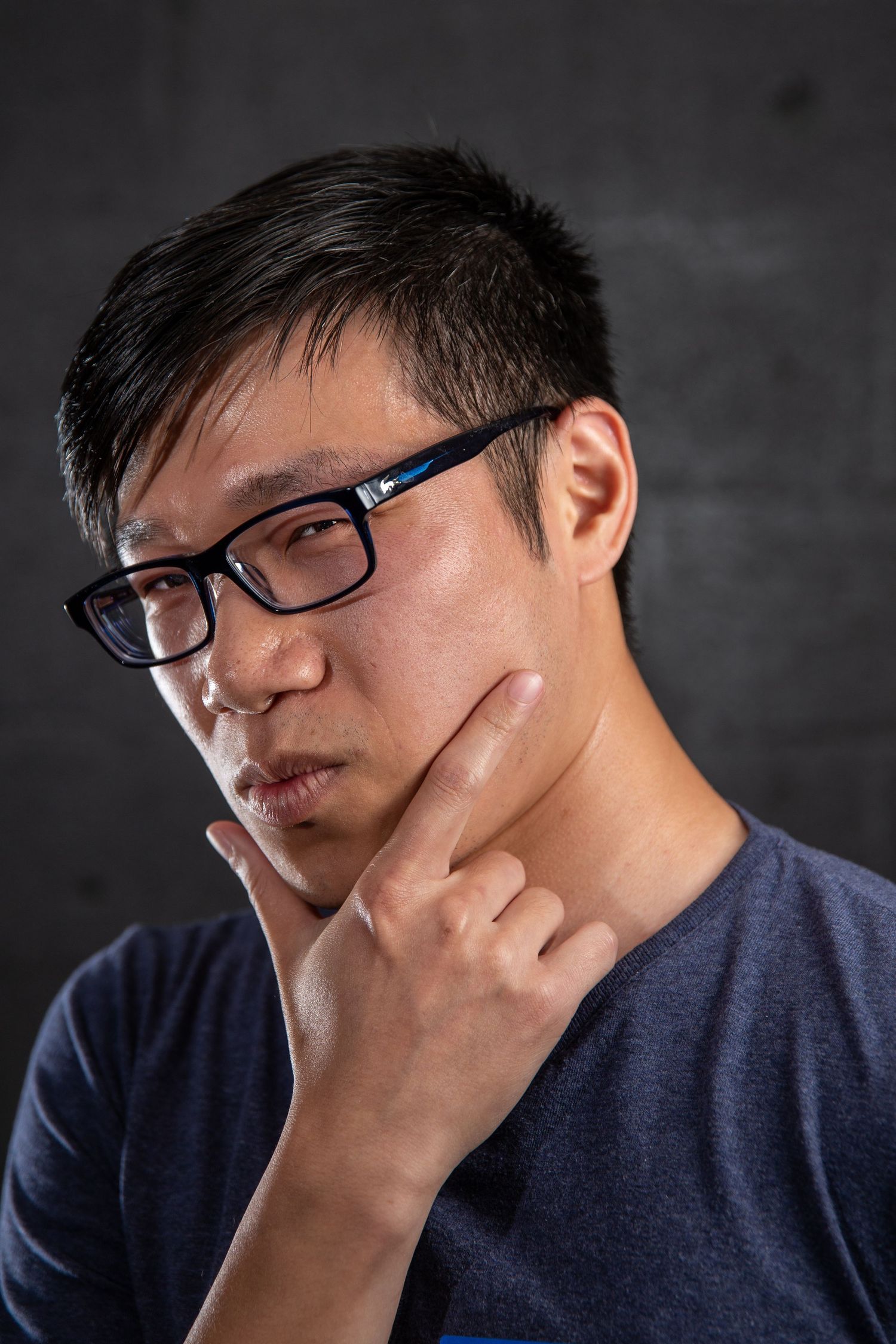 Thoughtful Asian man with glasses resting chin on hand against dark background.