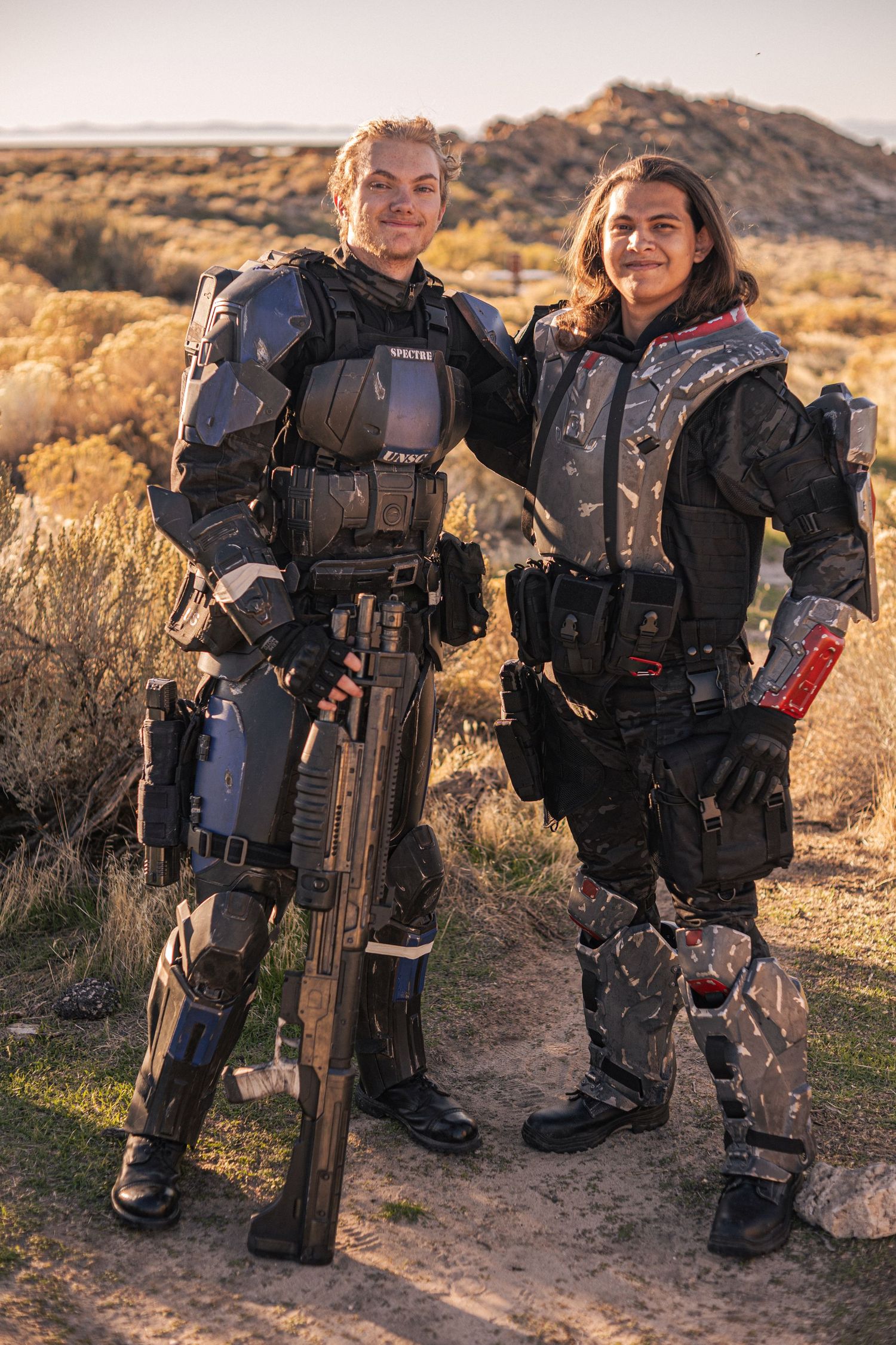 Two people posing in sci-fi combat armor costumes with large weapon prop in desert landscape.