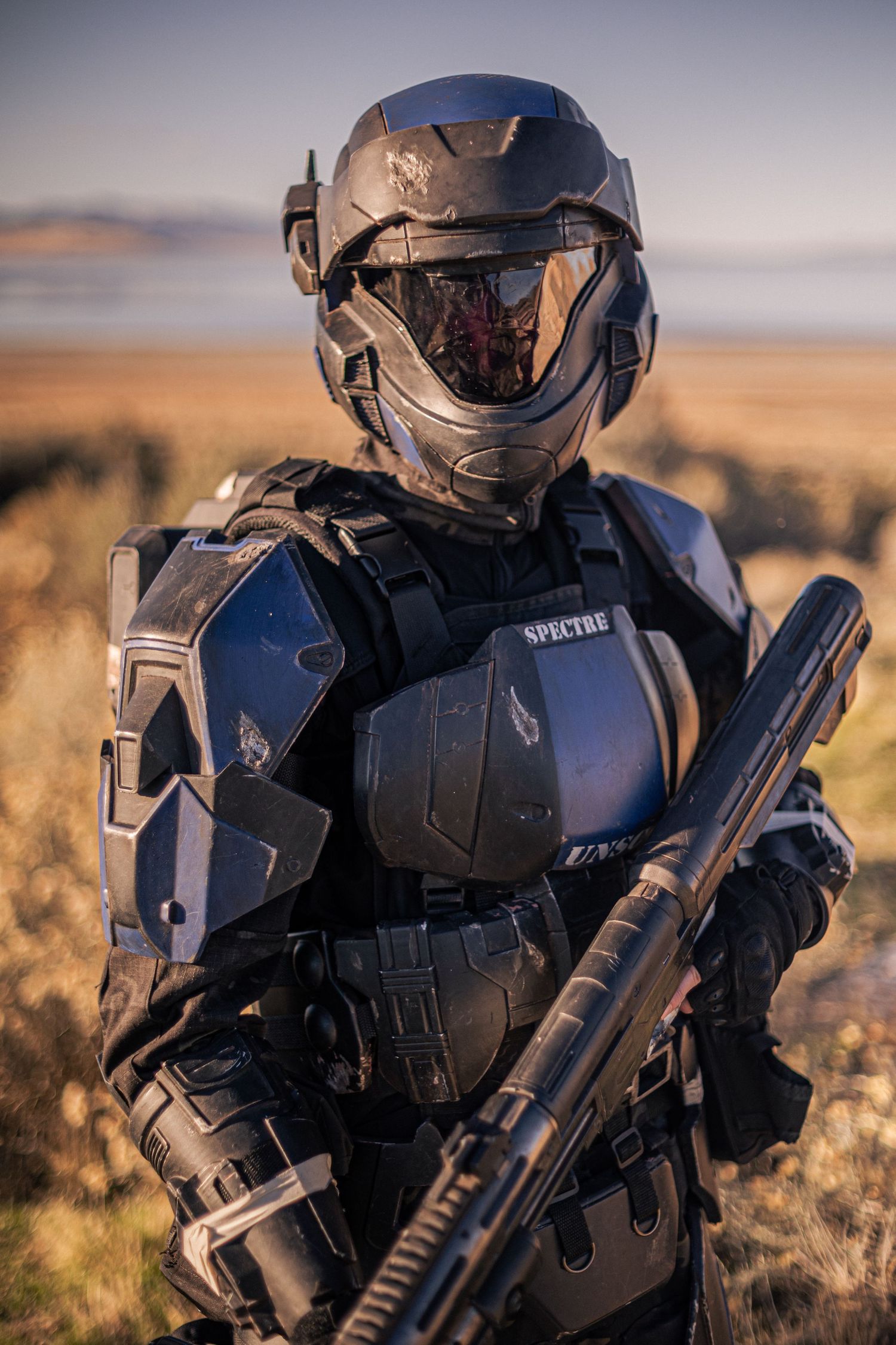 Cosplayer in black SPECTRE sci-fi military armor holding rifle in desert field.