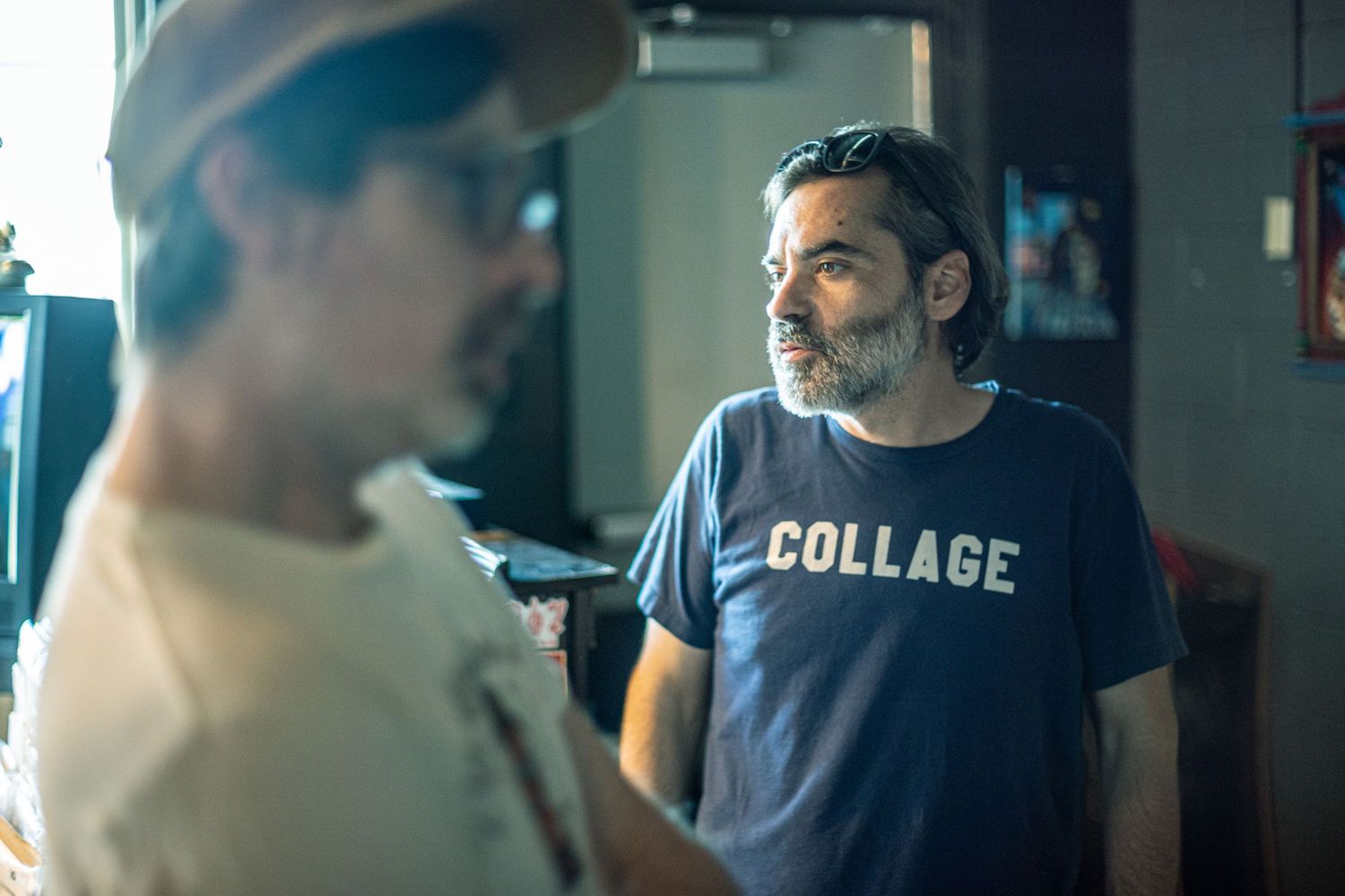 Two men having a conversation indoors, one wearing a navy blue shirt with the word COLLAGE printed on it.