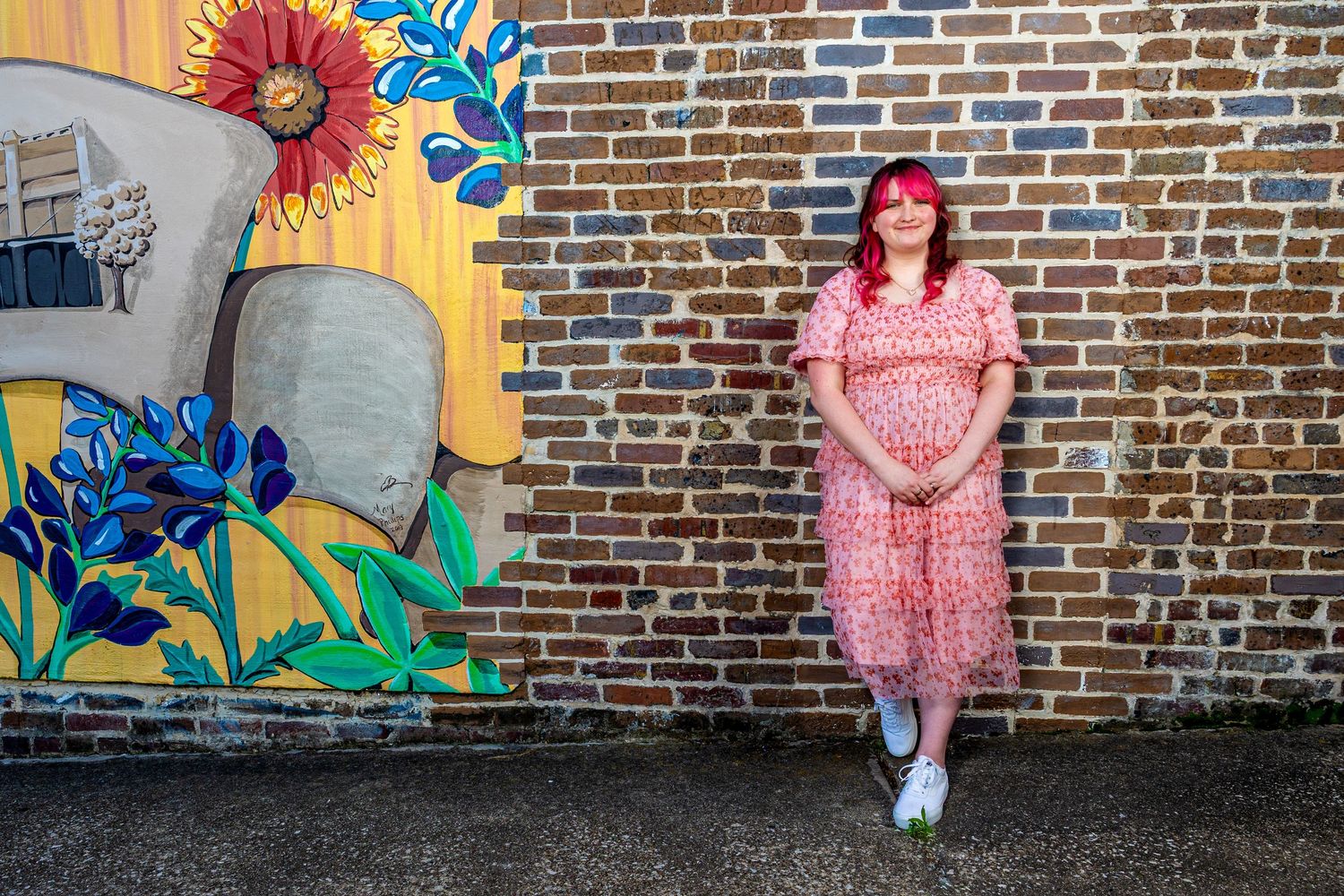 Pregnant woman in pink floral dress poses by brick wall with colorful mural featuring sunflower and blue flowers.