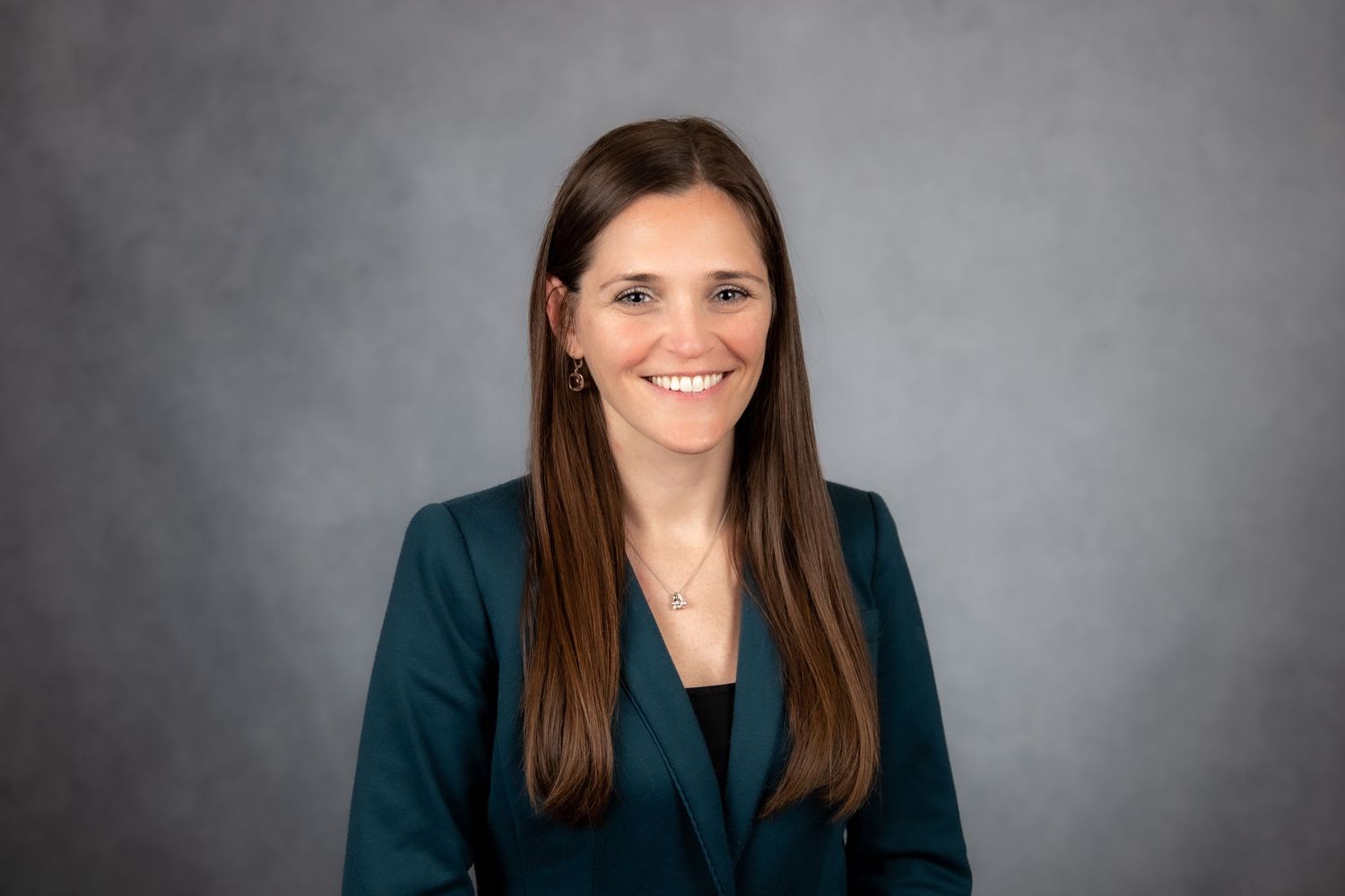 Professional headshot of a smiling woman with long brown hair wearing a teal blazer against a gray background.