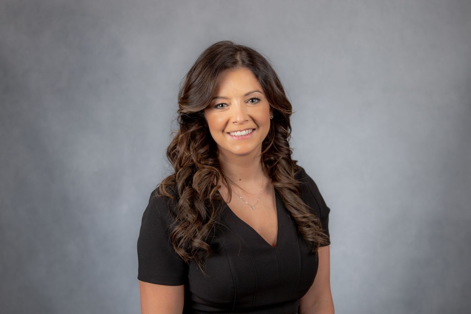 Smiling brunette woman in black dress poses for professional headshot on gray background.