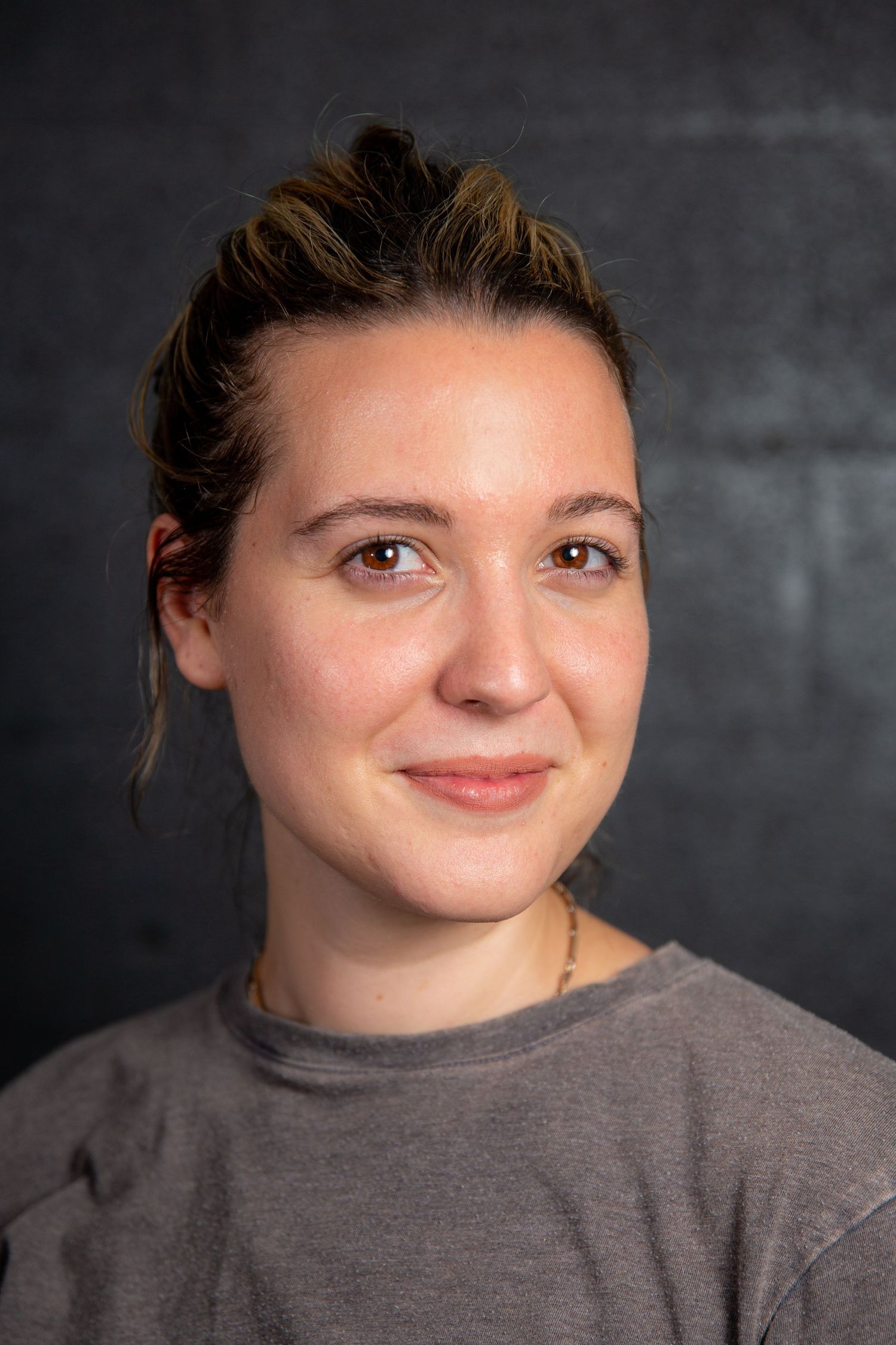 Young woman with brown hair in updo smiles softly in gray t-shirt against dark backdrop.