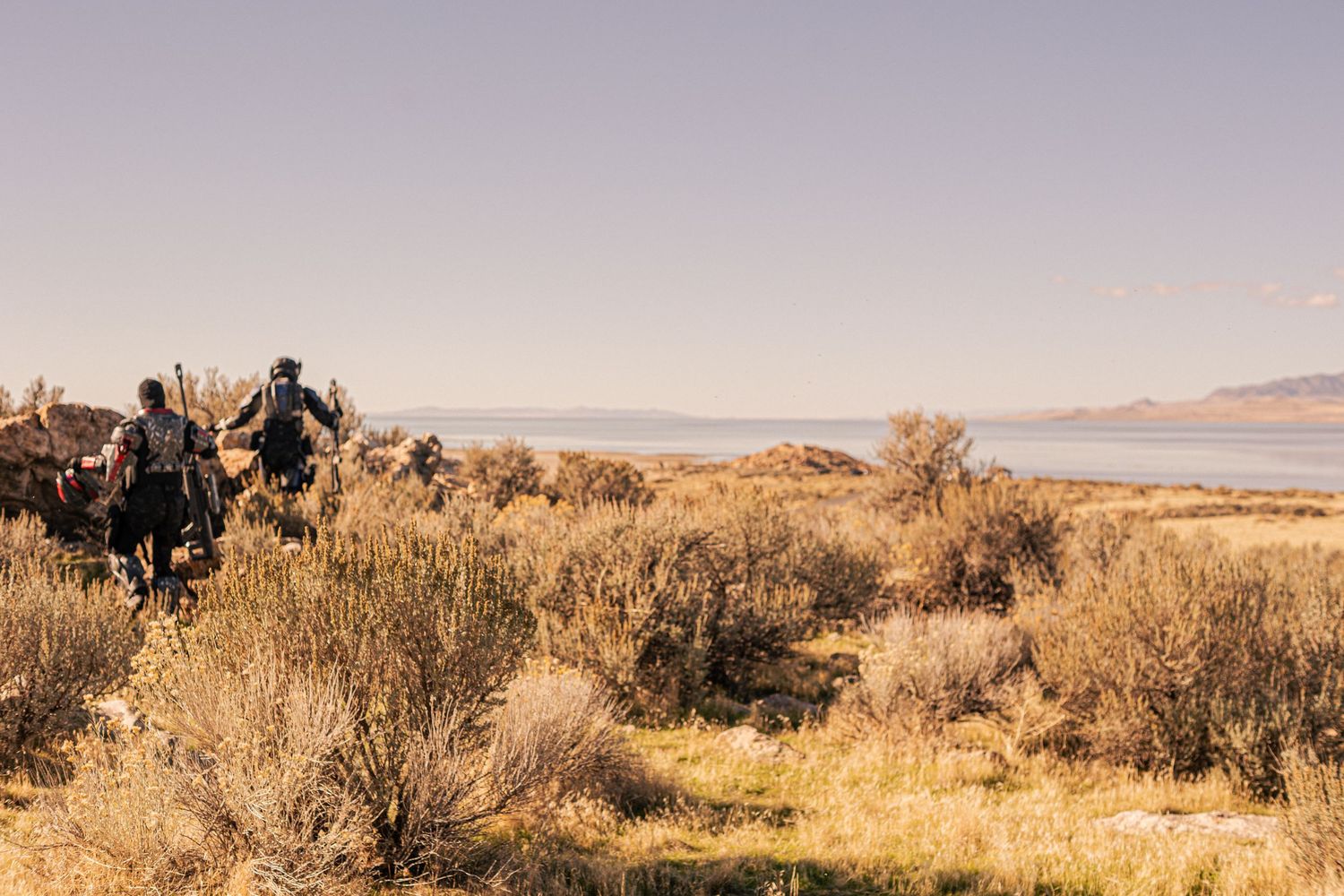 Hikers trekking through arid desert scrubland with a vast lake and clear sky in the background.