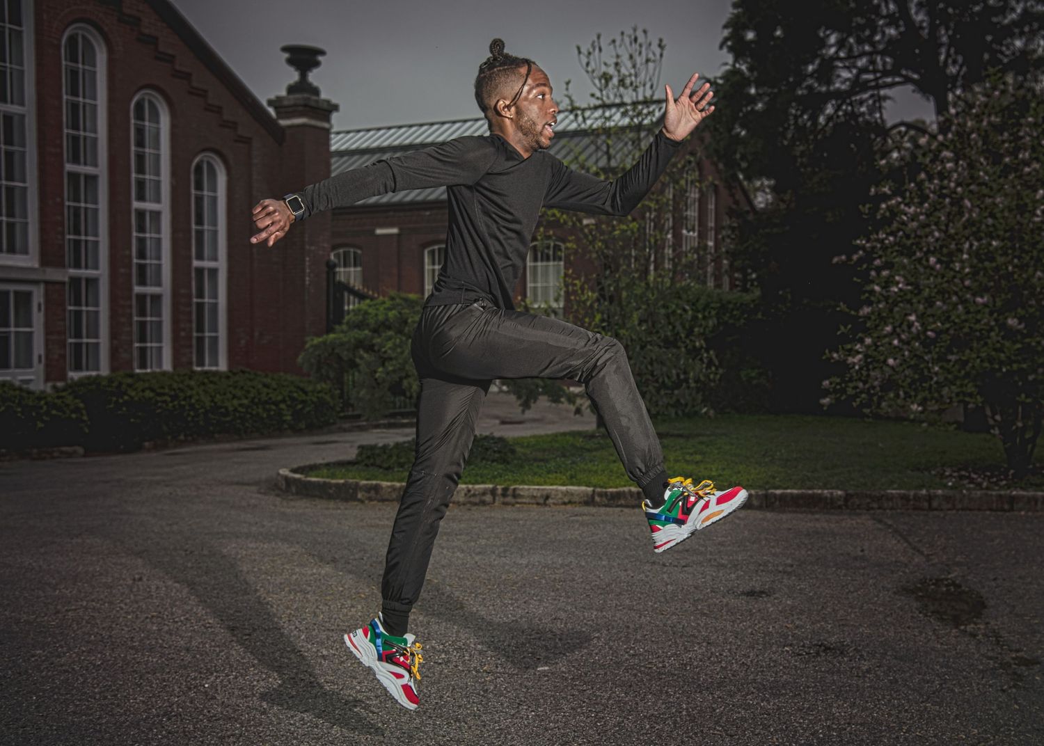 A person jumping mid-air wearing colorful sneakers and black outfit in front of a brick building.
