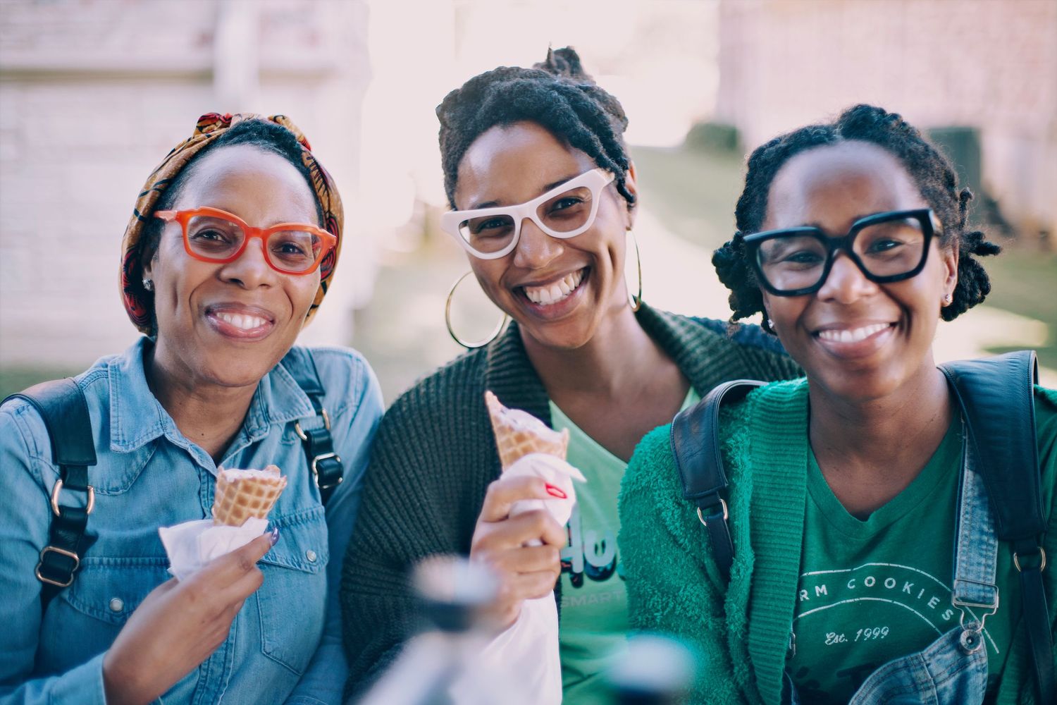 Three smiling women holding ice cream cones outdoors, wearing colorful glasses, enjoying a sunny day together.