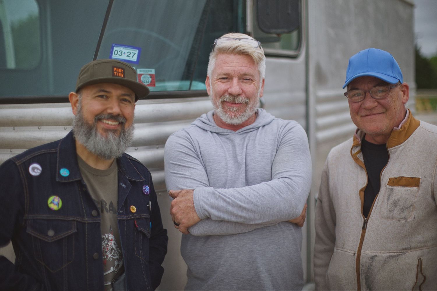 Three men smiling together outdoors in front of a vintage silver trailer on cloudy day.