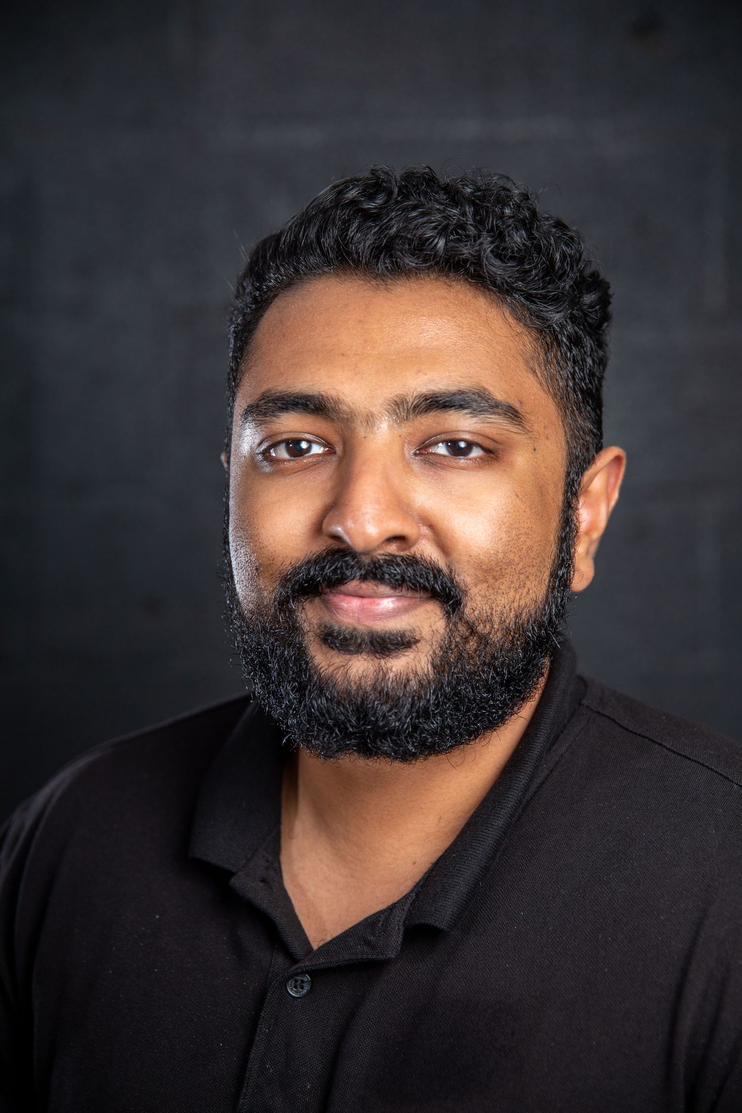Young man with black curly hair and beard smiles in black shirt against dark background.