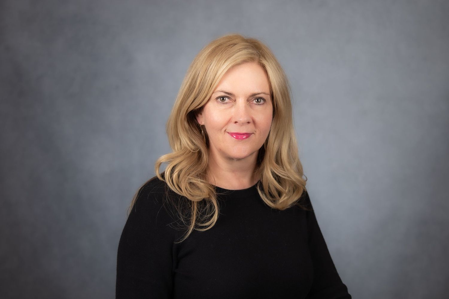 Professional blonde woman in black top smiles for corporate headshot on gray background.