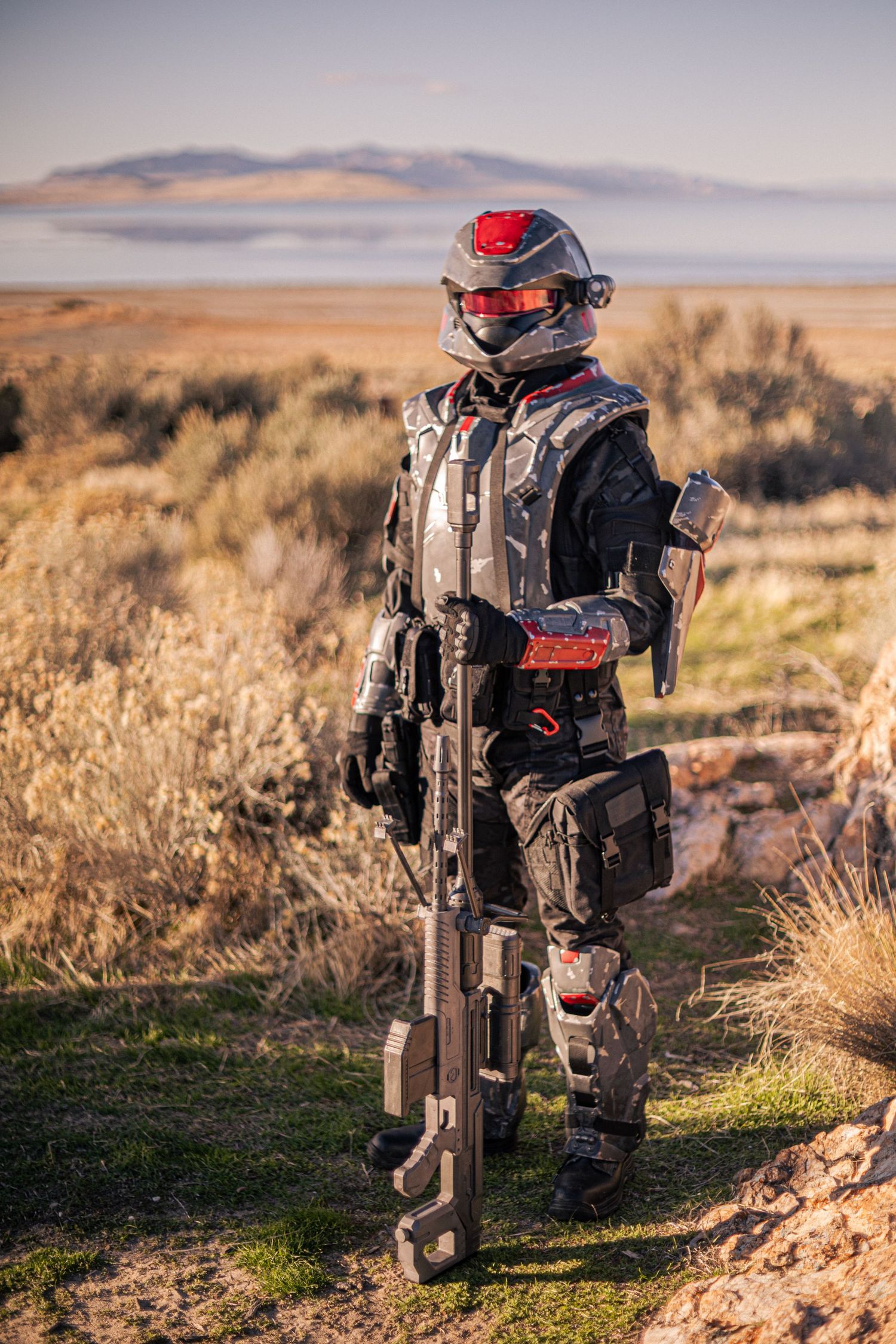 Cosplayer in red and black futuristic military armor holding a sci-fi rifle in a desert shrubland setting.
