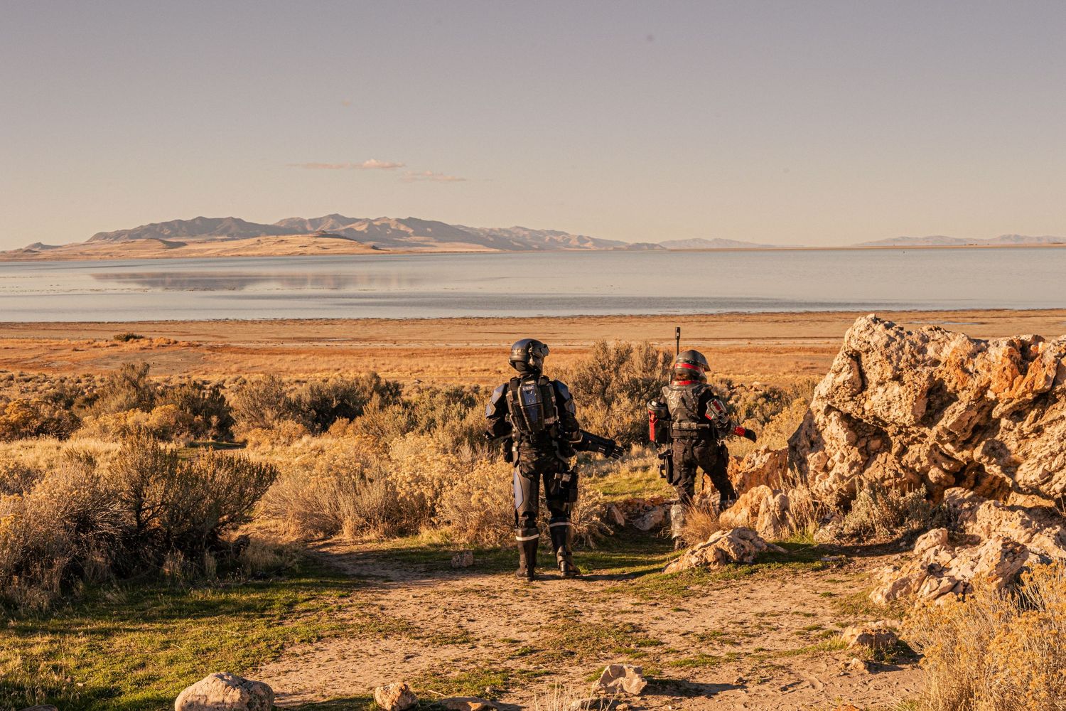Two cosplayers in sci-fi armor overlook a vast desert lake surrounded by golden shrubland.