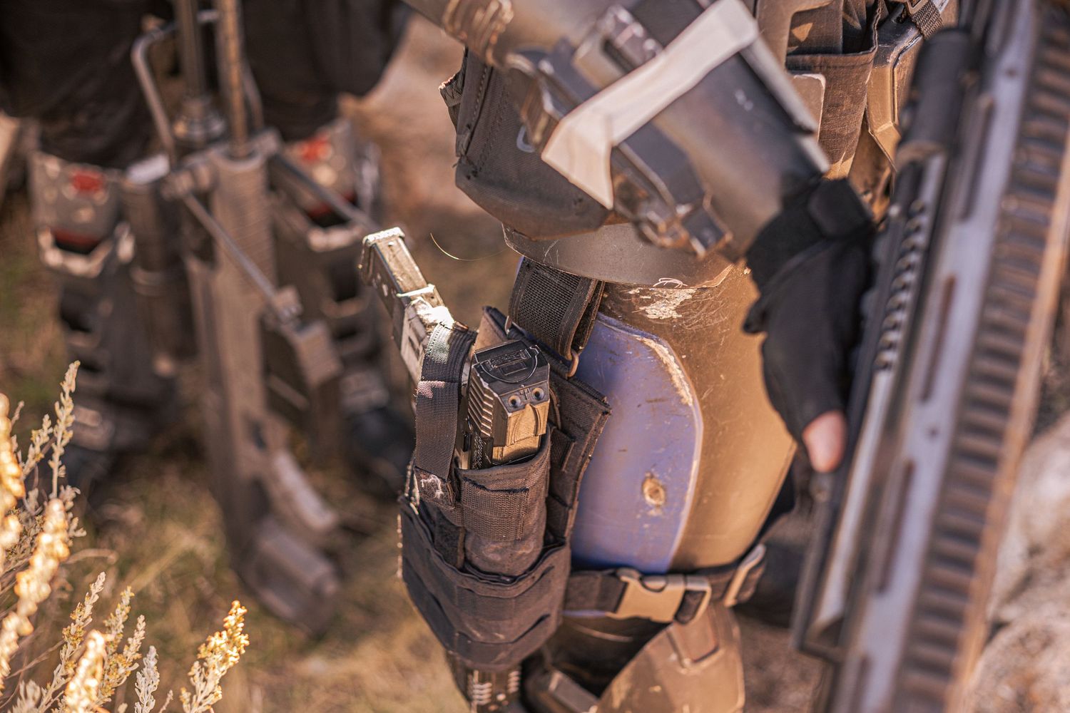 Close-up of a tactical holster with a pistol attached to armored military gear outdoors.