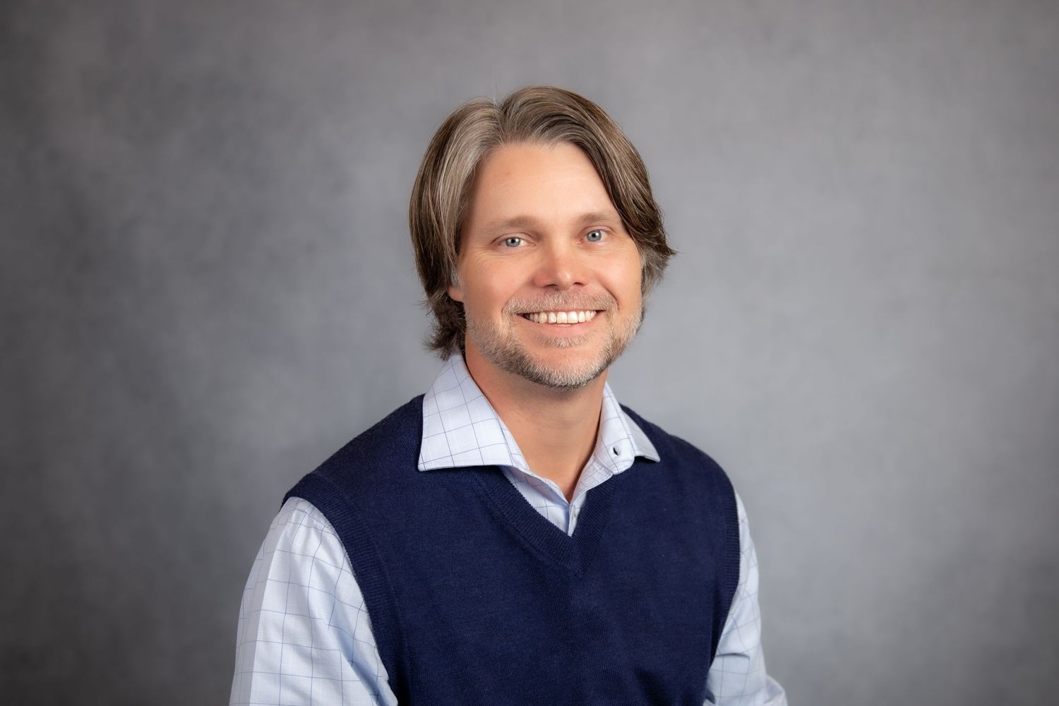 Professional headshot of a smiling man with medium-length brown hair wearing a navy vest and light blue collared shirt.
