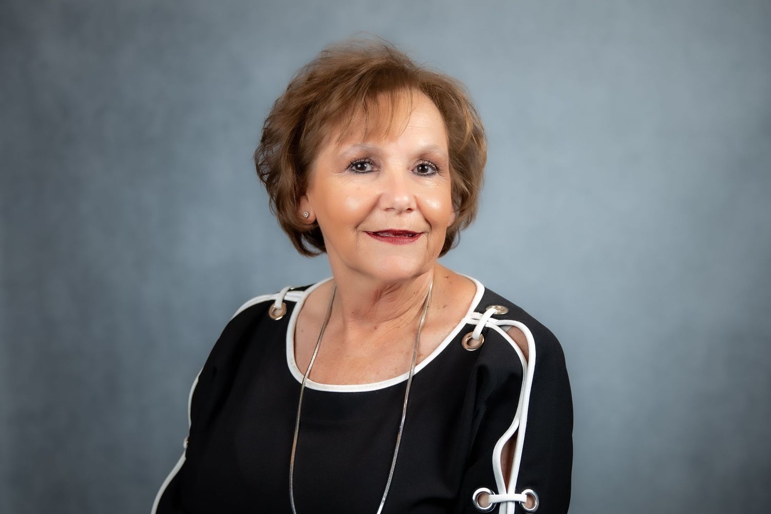 Older woman smiling in a black top with white trim and a silver necklace against a gray background.