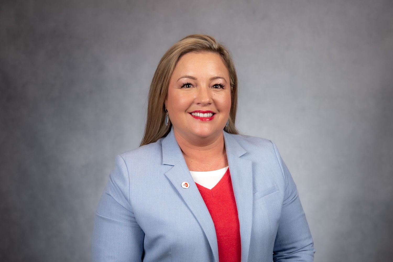 Smiling woman with blonde hair wearing a light blue blazer and red top, professional headshot on gray background.
