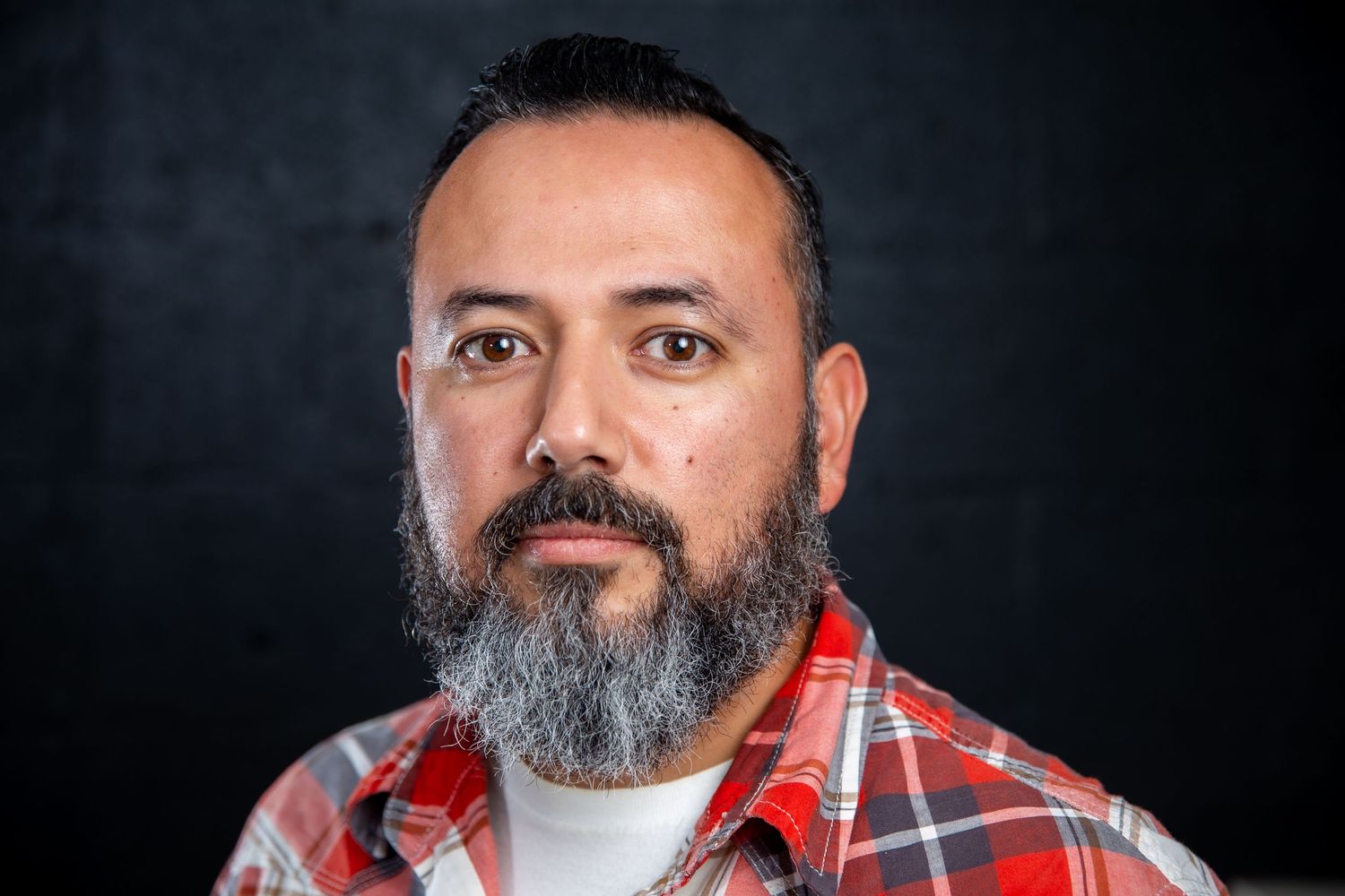 Bearded Hispanic man in red plaid shirt poses for professional headshot against dark background.