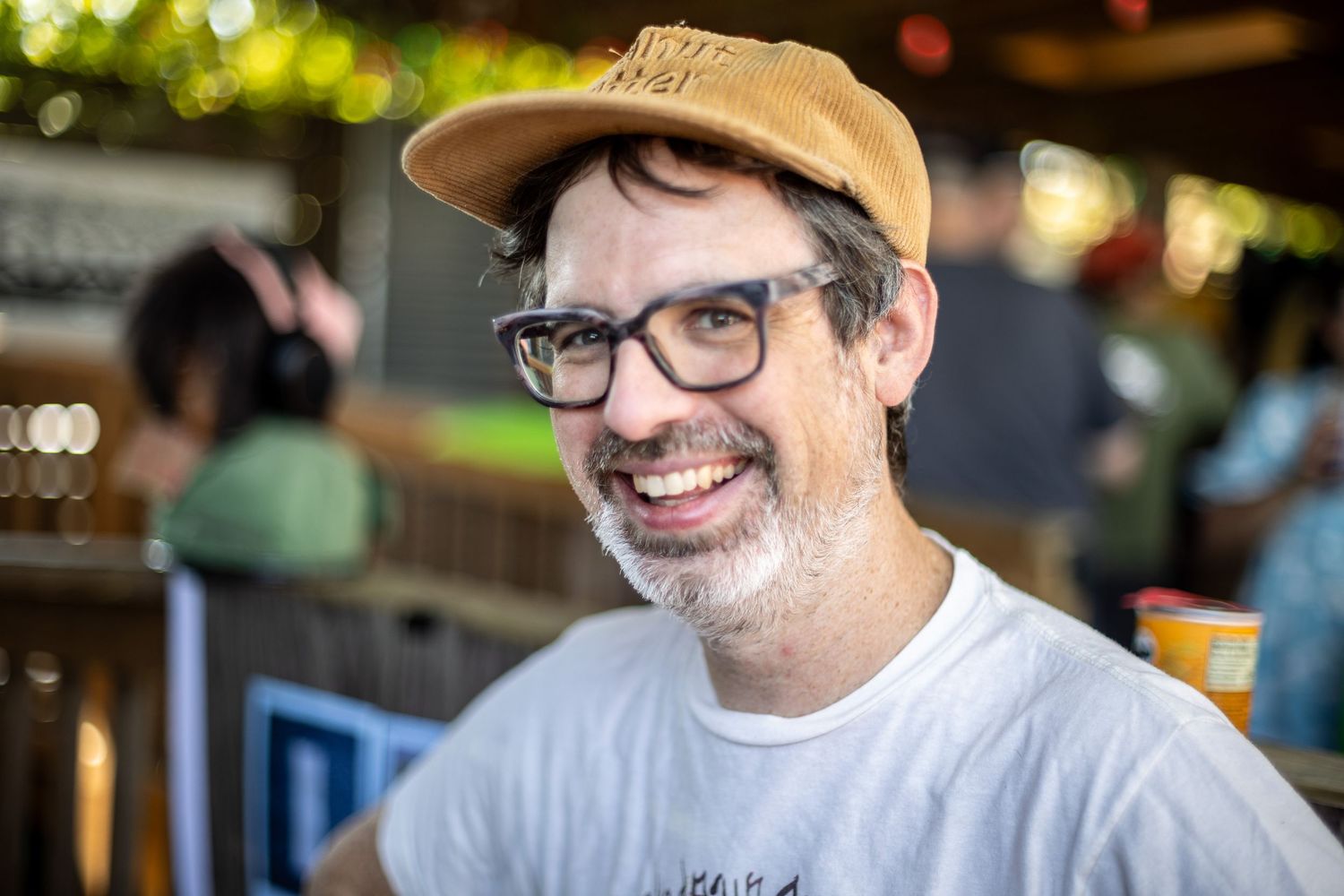 Smiling man with glasses and a tan cap enjoying drinks at an outdoor bar or restaurant patio.