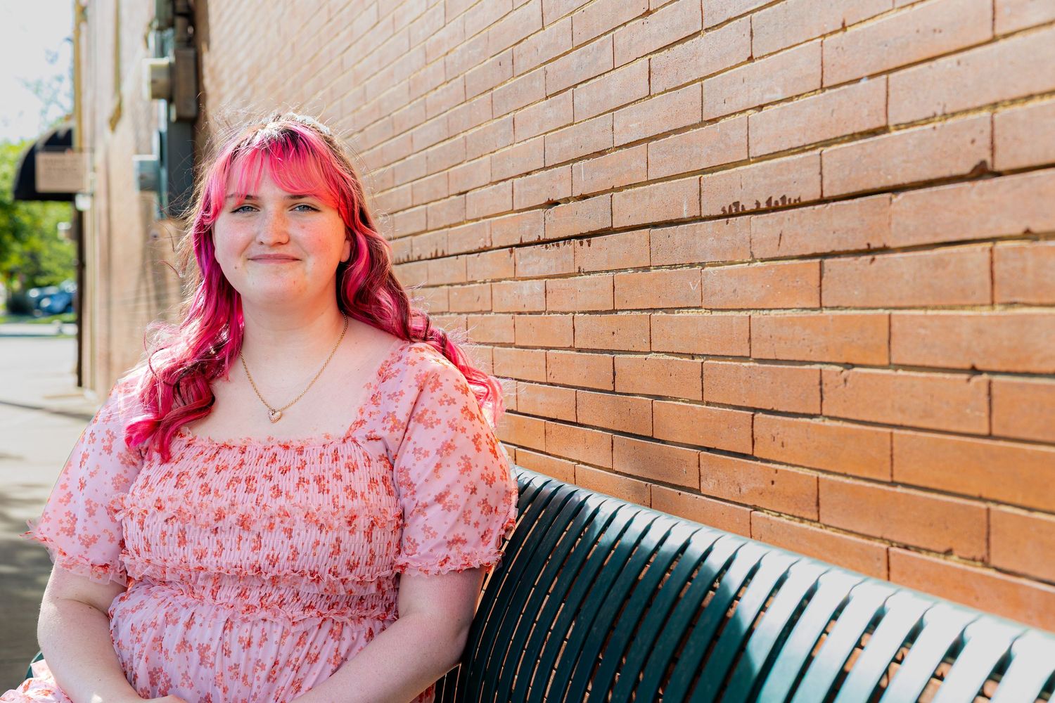 Young woman with pink hair in floral dress sitting on bench against red brick wall outdoors.