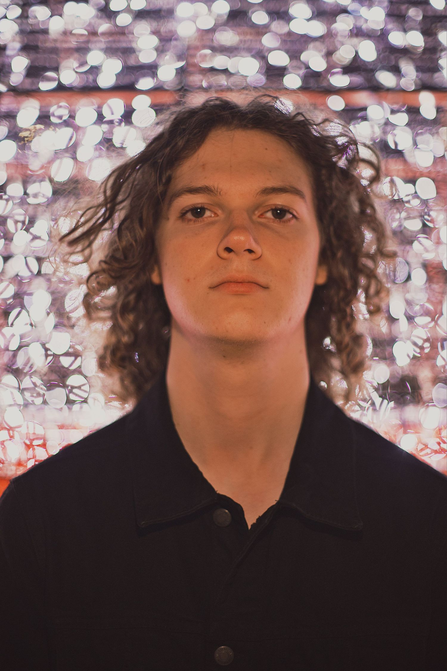Young man with curly hair wearing a black polo shirt posed against a bokeh light background.