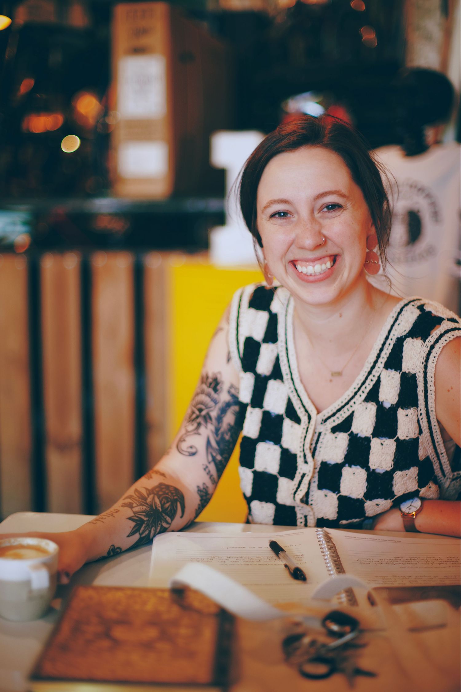Smiling woman with tattoos wearing a checkered top sits at a cafe table with coffee and an open notebook.
