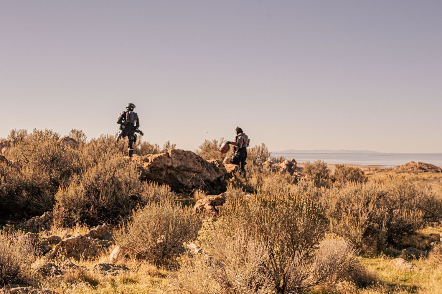 Two hikers traverse a rocky desert landscape with sagebrush under a clear sky.