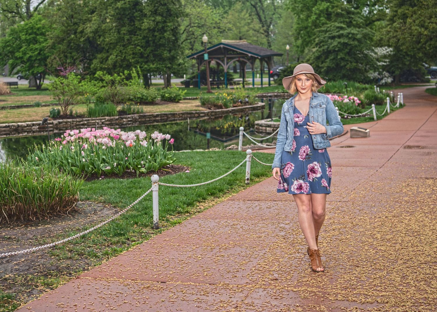 Woman in floral dress, denim jacket, and hat walking along a garden path with pink flowers and a gazebo.