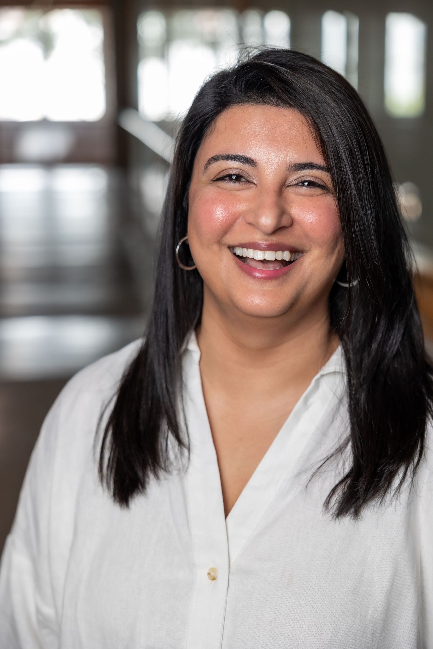 Smiling South Asian woman with black hair wearing white blouse and gold hoop earrings.