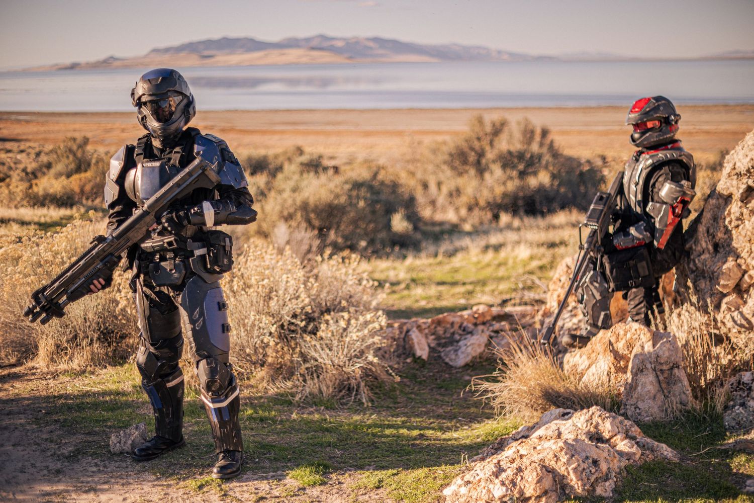Two cosplayers in detailed sci-fi combat armor pose with weapons in a desert shrubland setting.