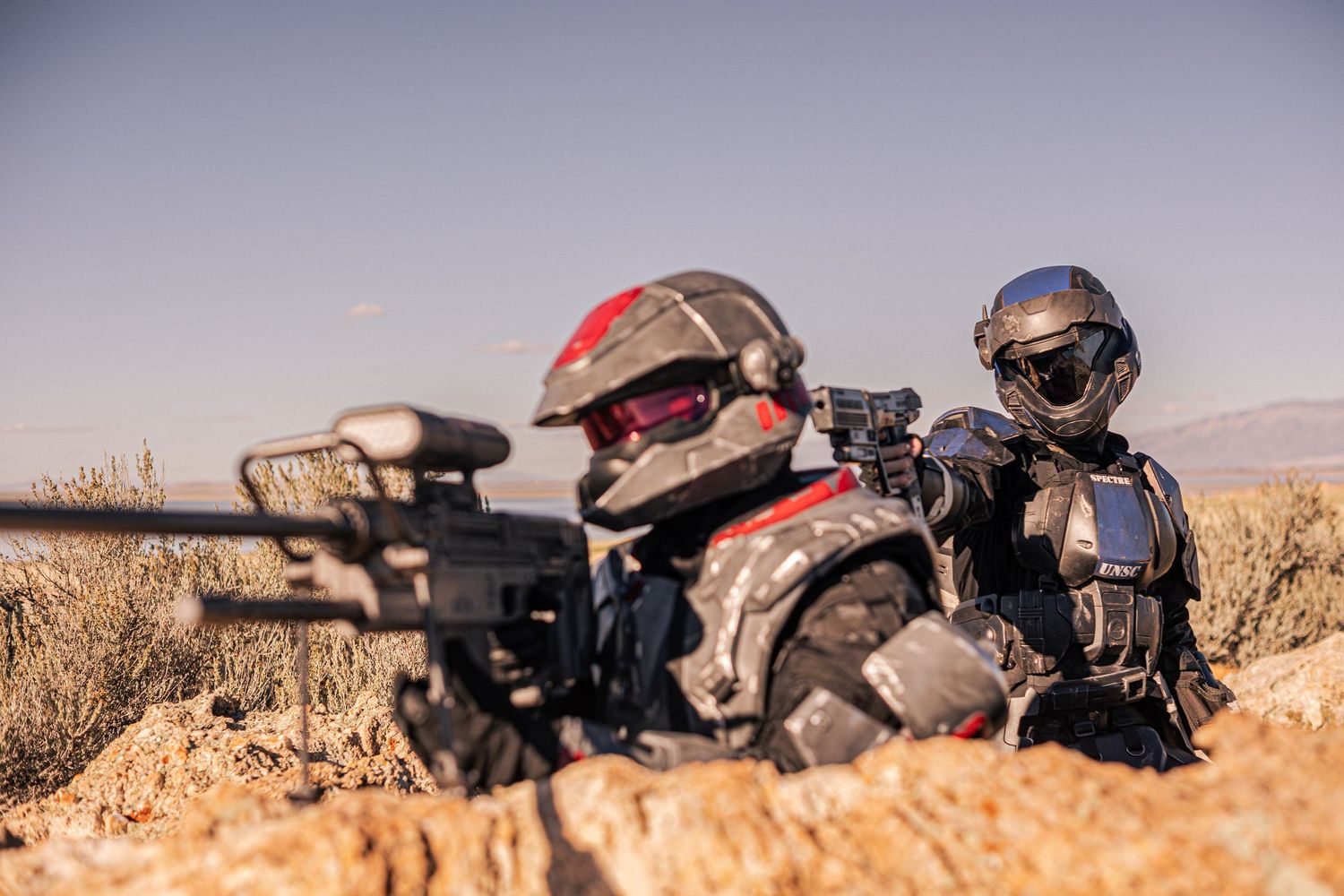 Two futuristic soldiers in sci-fi cosplay armor aiming sniper rifle in desert.