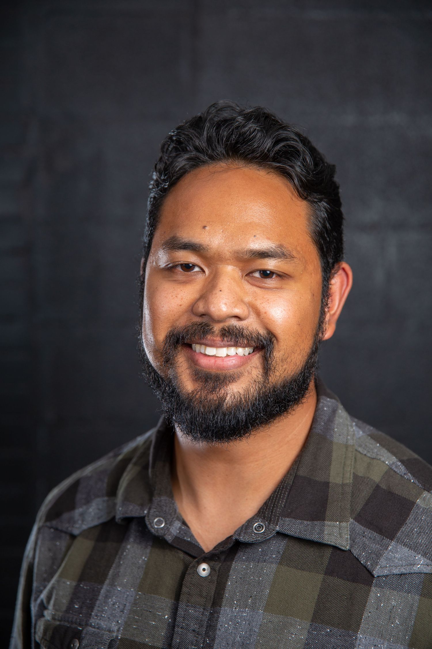 Smiling young man with beard wearing plaid shirt against dark background.