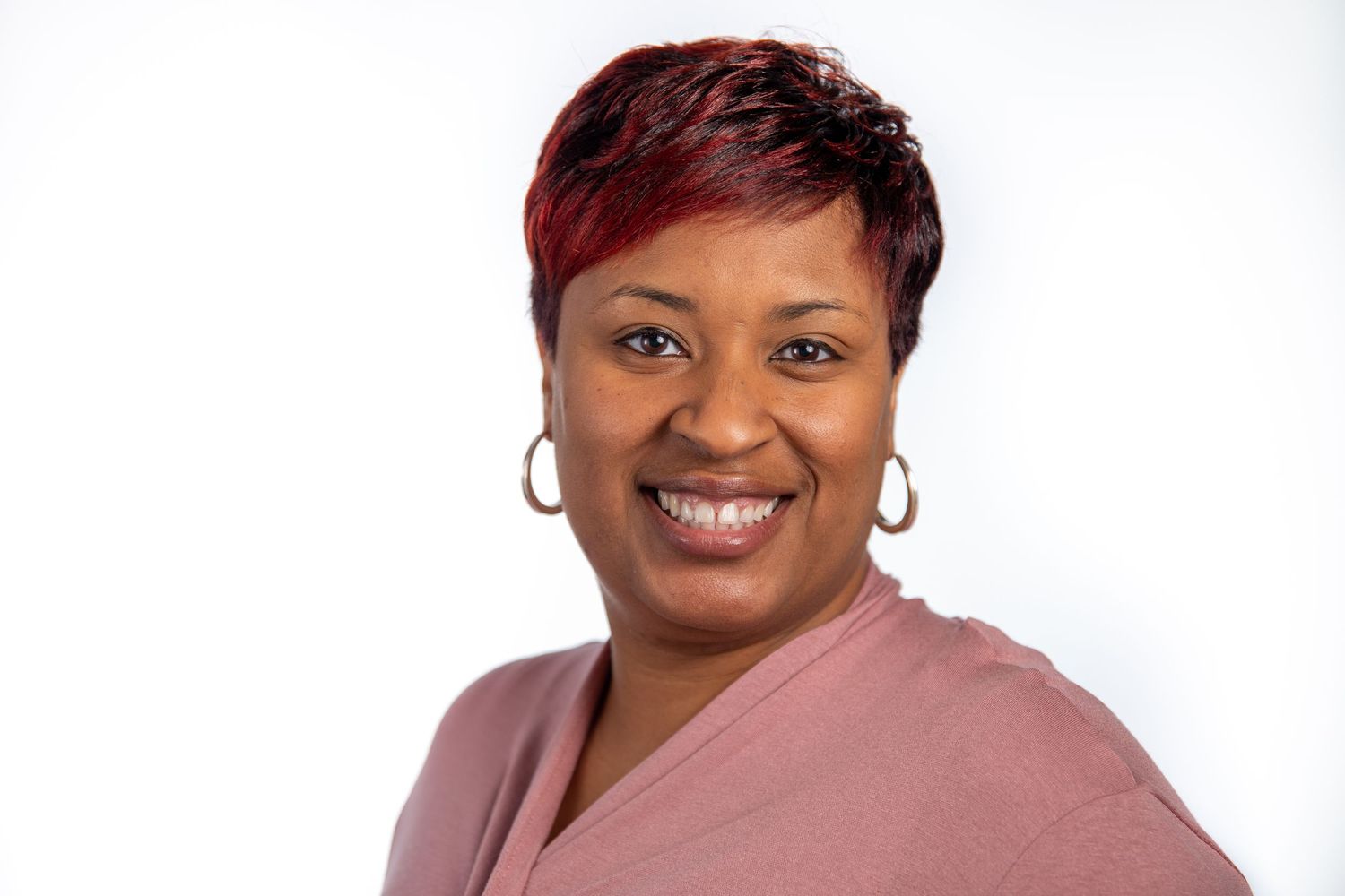 Smiling woman with short red hair wearing a mauve top and gold hoop earrings on white background.