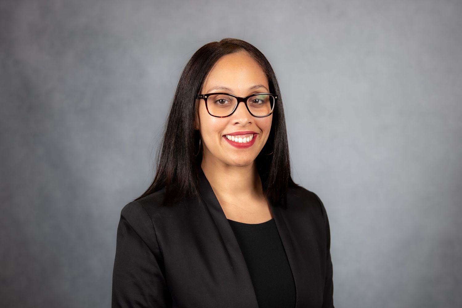 Professional headshot of a smiling woman with glasses and straight black hair in a black blazer on gray background.