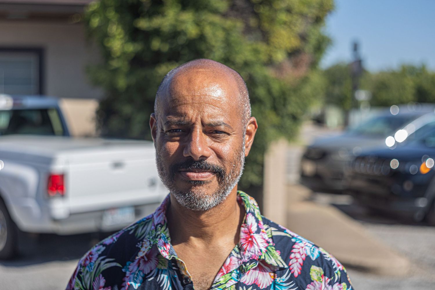 Middle-aged man with beard wearing floral shirt standing outdoors near parked vehicles.