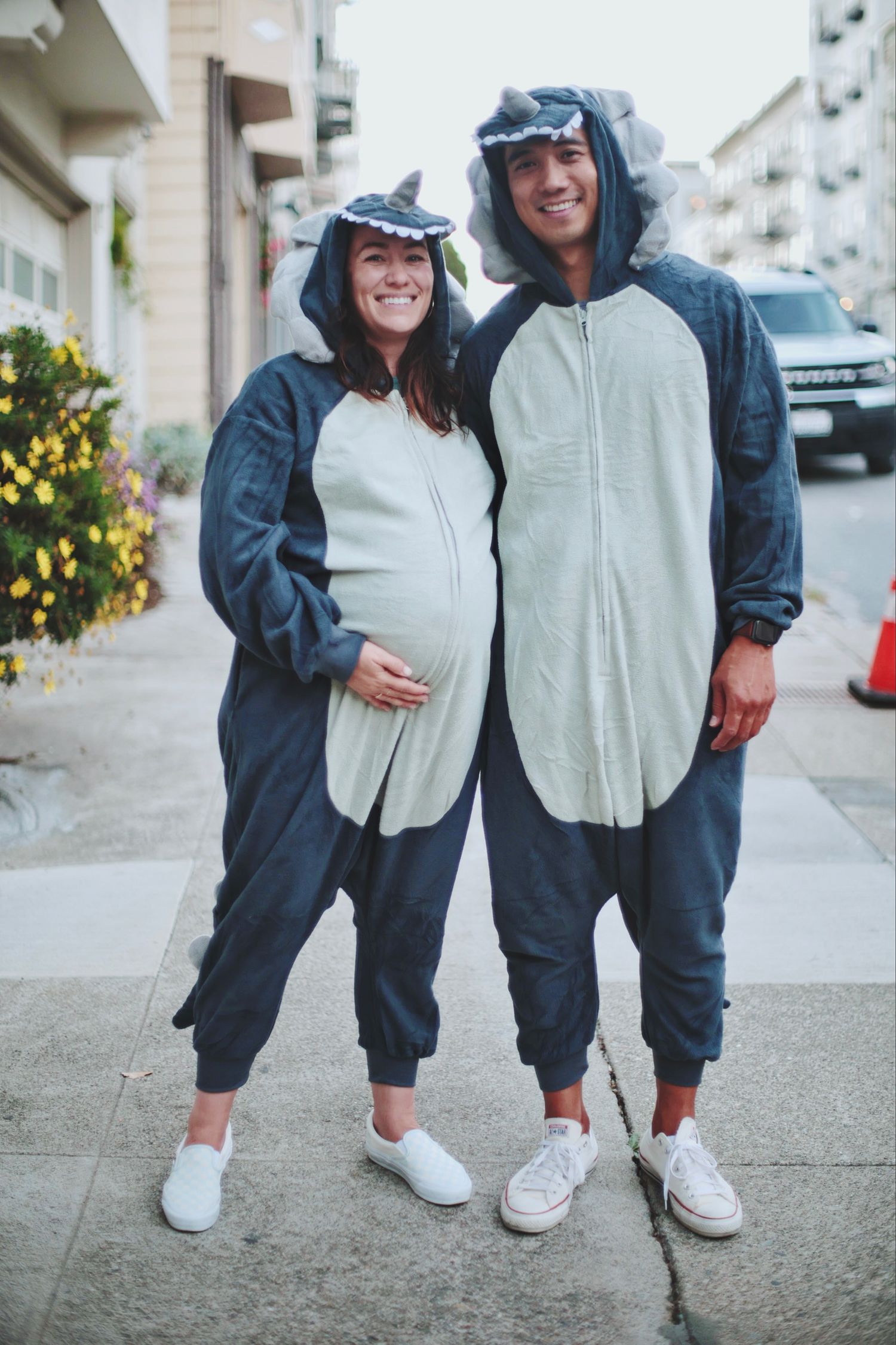 Pregnant couple in matching shark onesie costumes smiling on a sidewalk.