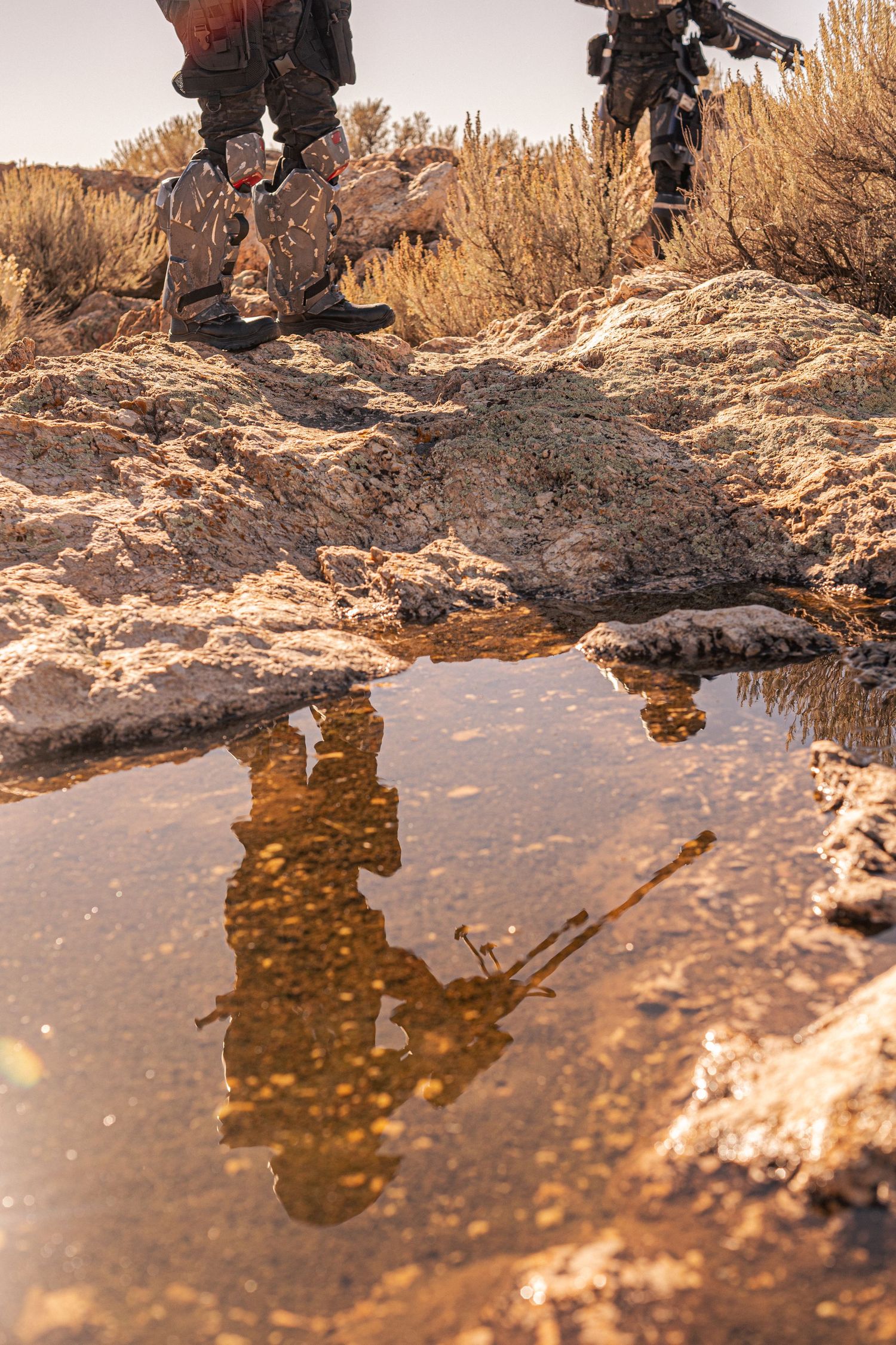 Reflection of armed soldiers in a desert puddle surrounded by rocky terrain and dry brush.