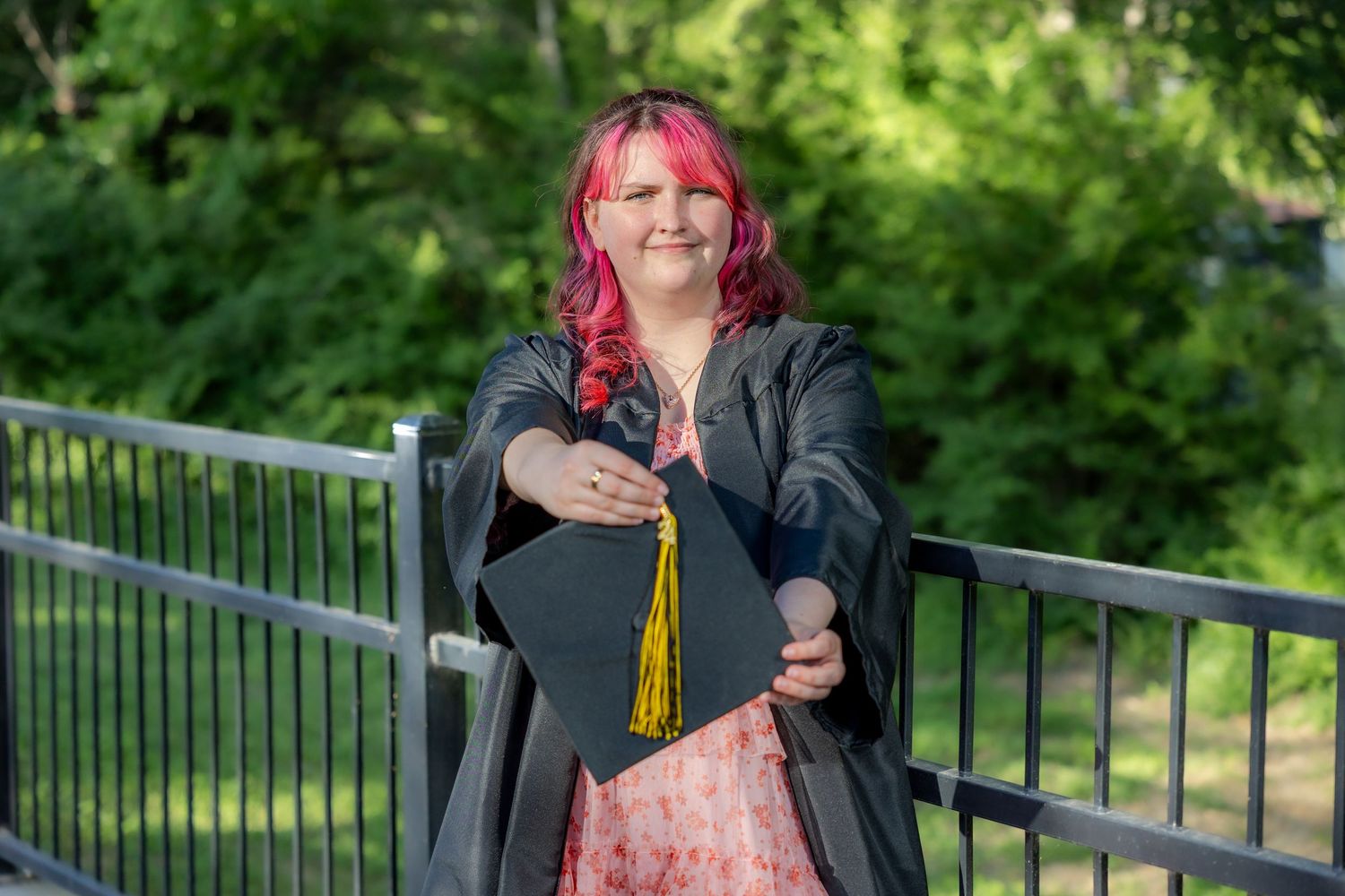 A graduate with pink hair holds cap and diploma wearing black gown on a bridge outdoors.