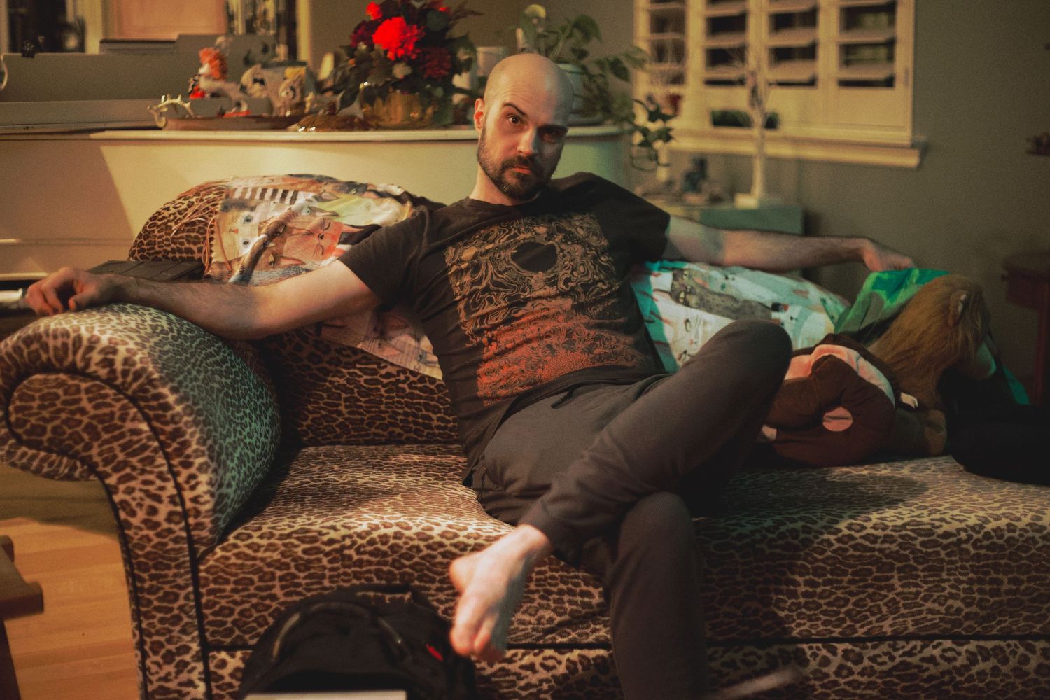 Bearded man relaxing on a leopard print sofa in a cozy living room wearing a graphic t-shirt.