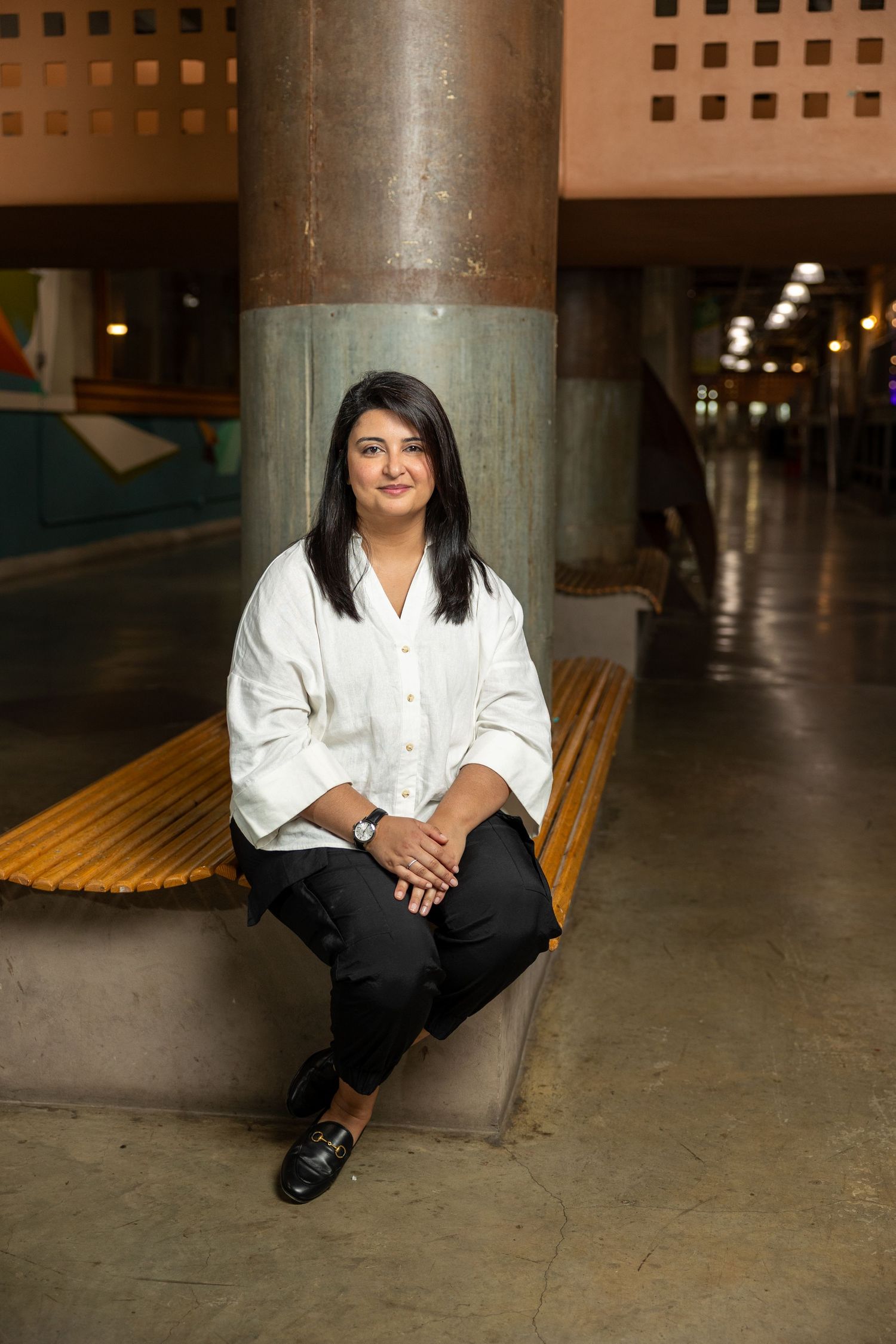 South Asian woman with black hair in white blouse sitting on bench in modern architectural space.