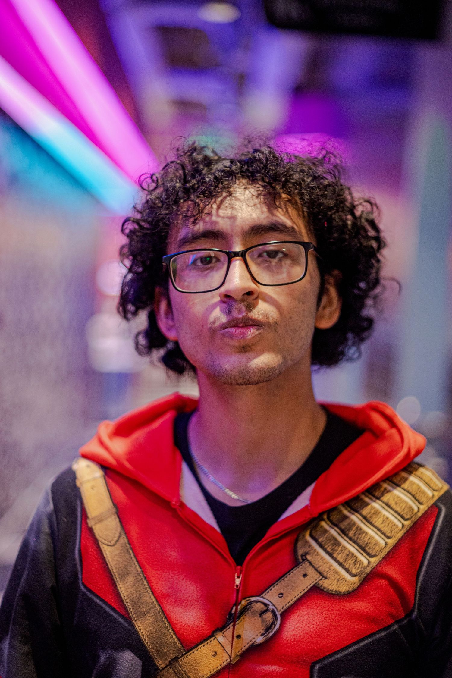 Young man with curly hair and glasses wearing a red superhero costume at a gaming event.