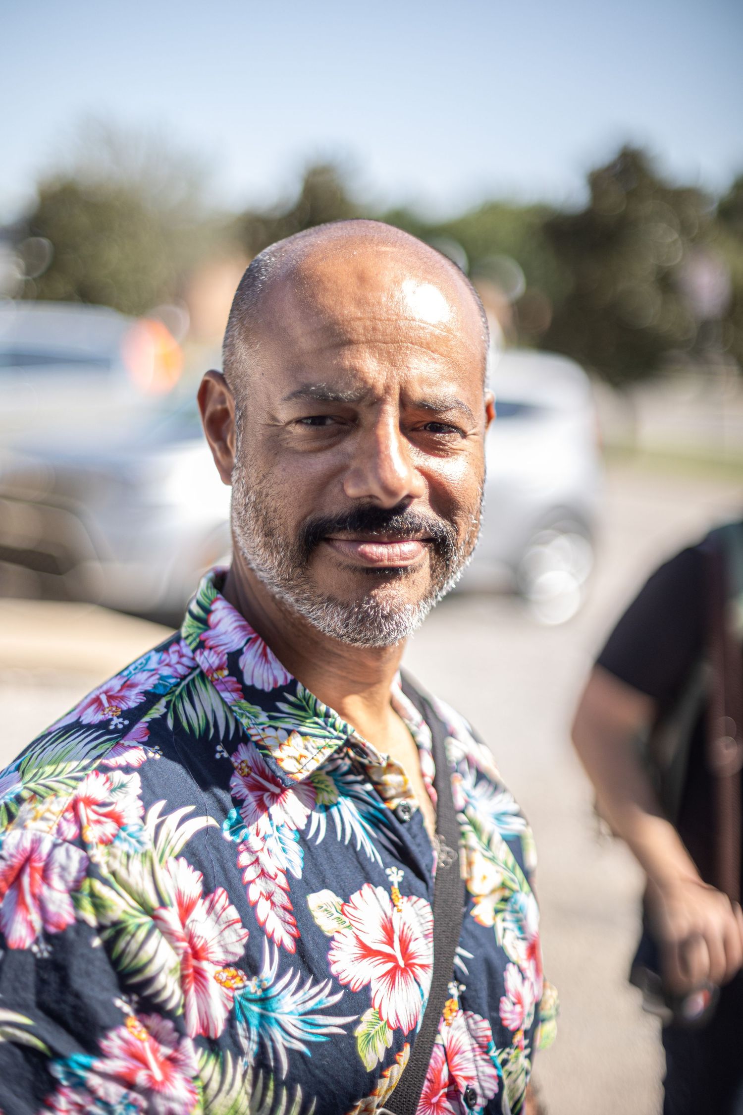 Bald man with gray beard smiles wearing a colorful floral Hawaiian shirt outdoors on sunny day.