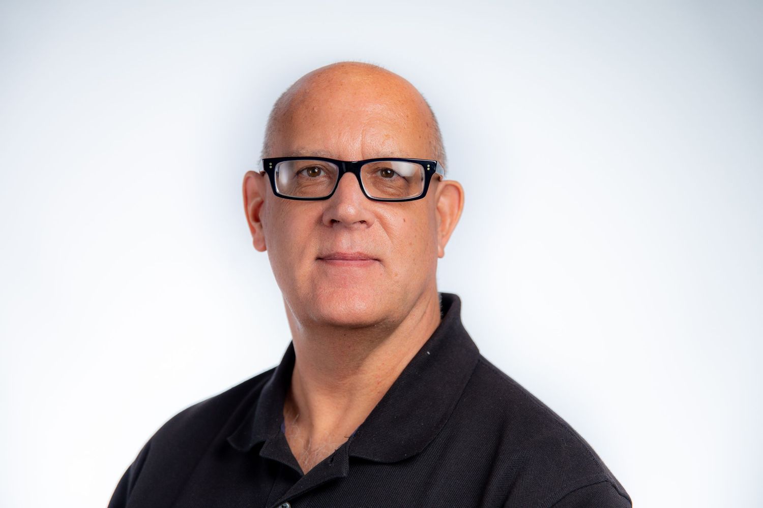 Bald man wearing black-rimmed glasses and a black polo shirt poses for a professional headshot.