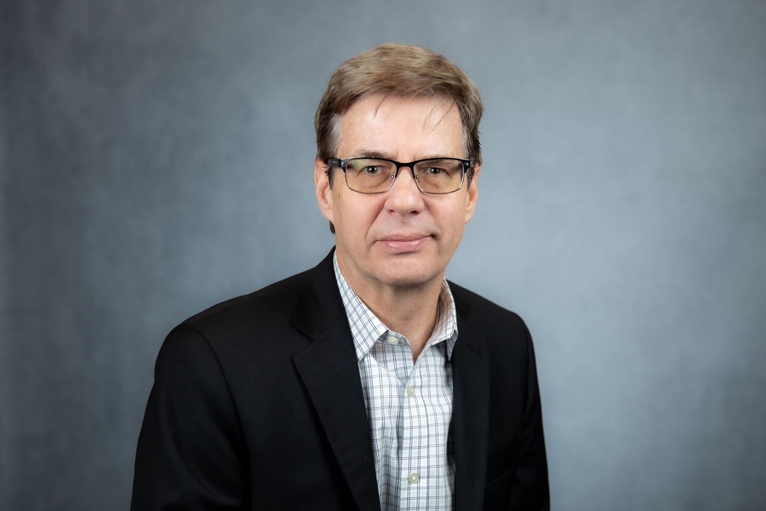 Professional older man wearing glasses and black blazer over checkered shirt on gray background.