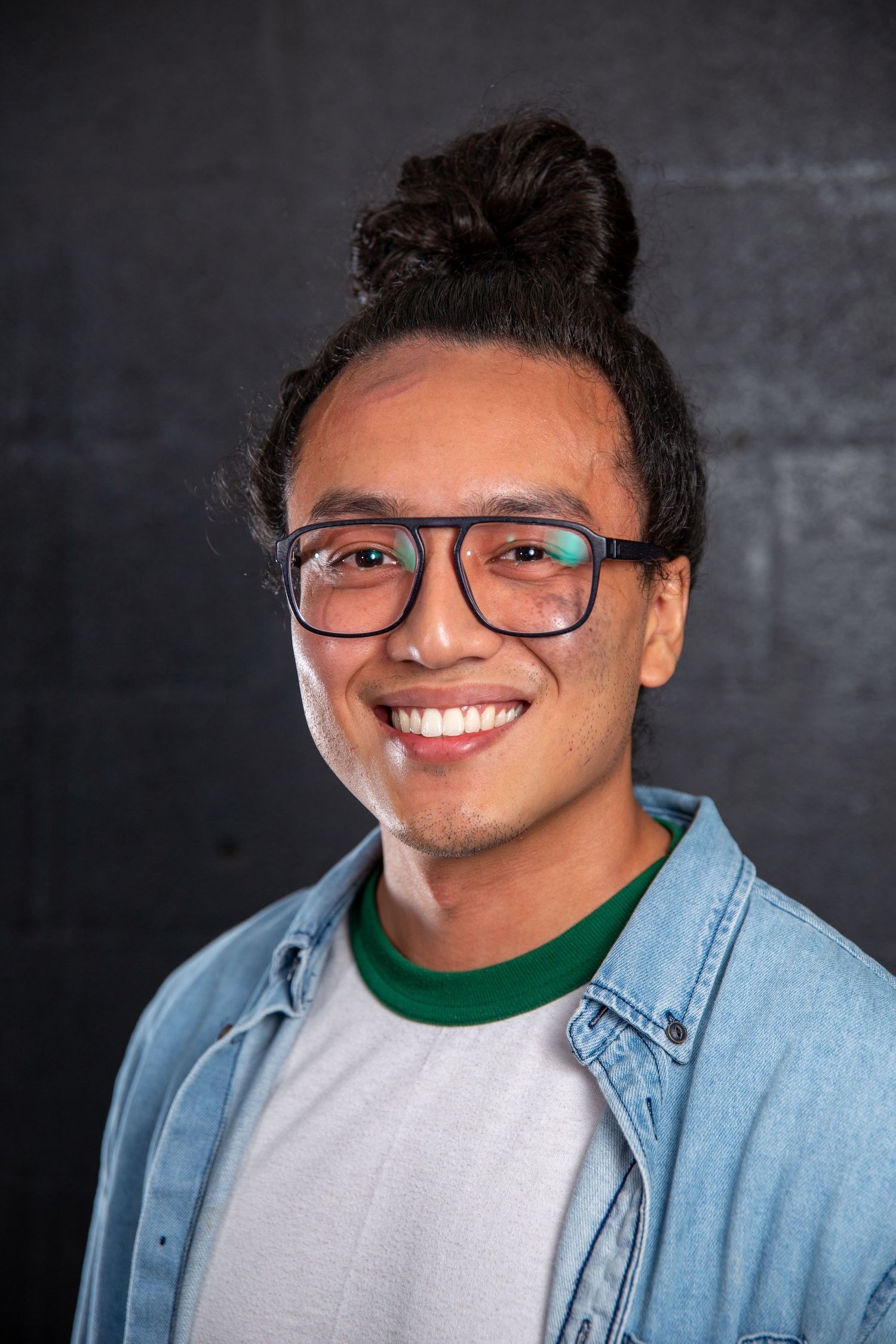 Smiling young Asian man with hair bun and glasses wearing a denim shirt over a white tee.