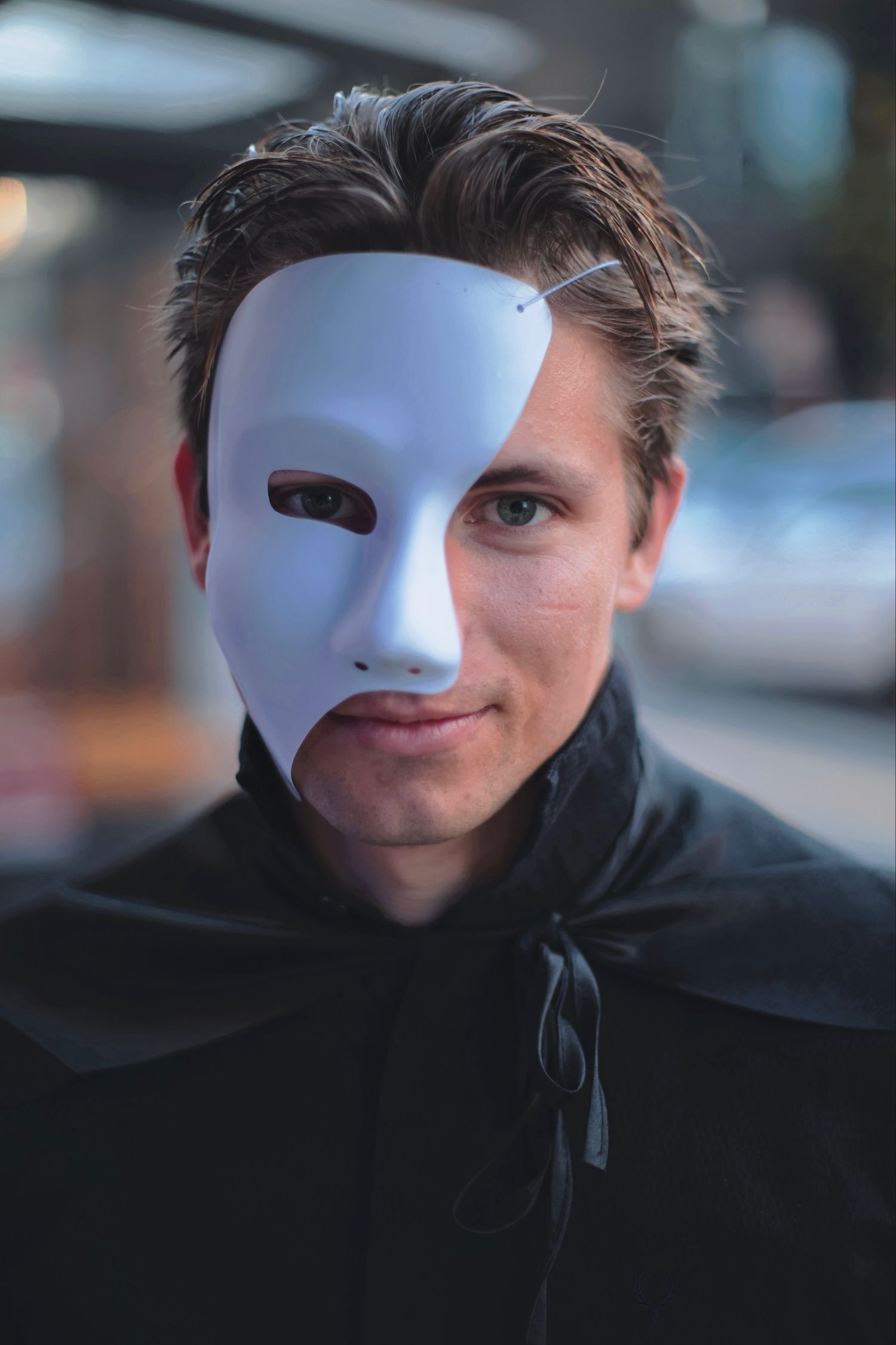 Young man wearing half white phantom mask and black cape in dramatic urban portrait.